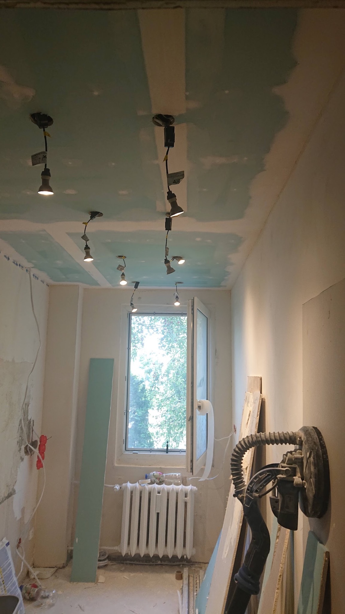 Wnętrze w trakcie prac wykończeniowych: szpachlowana sufit z zamontowanym oświetleniem szynowym, szlifierka do gipsu oparta o ścianę, widoczne płyty gipsowe i grzejnik pod oknem.