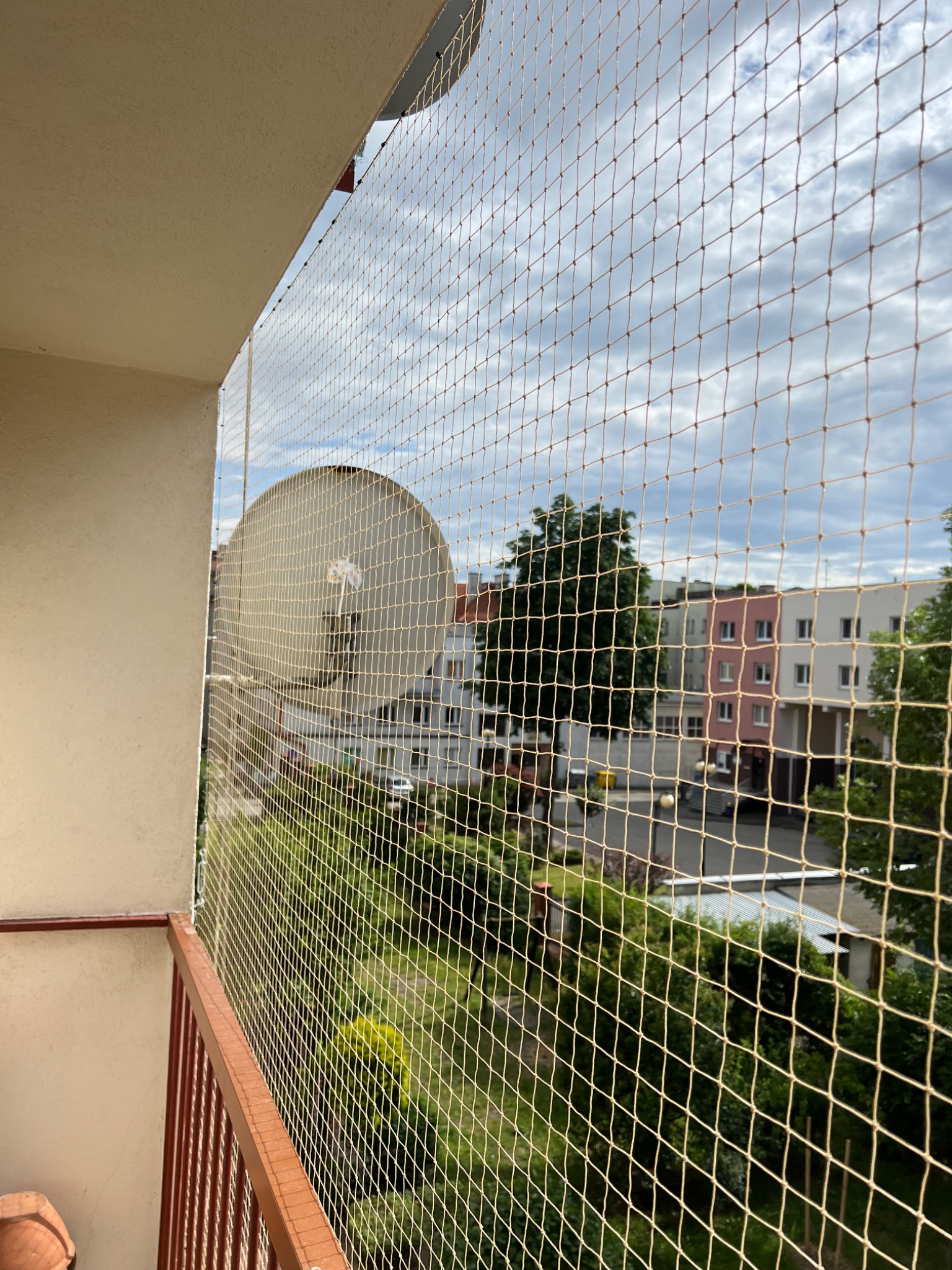 Beżowa siatka ochronna na balkonie z widokiem na zieleń i budynki mieszkalne, zamontowana na brązowej balustradzie.