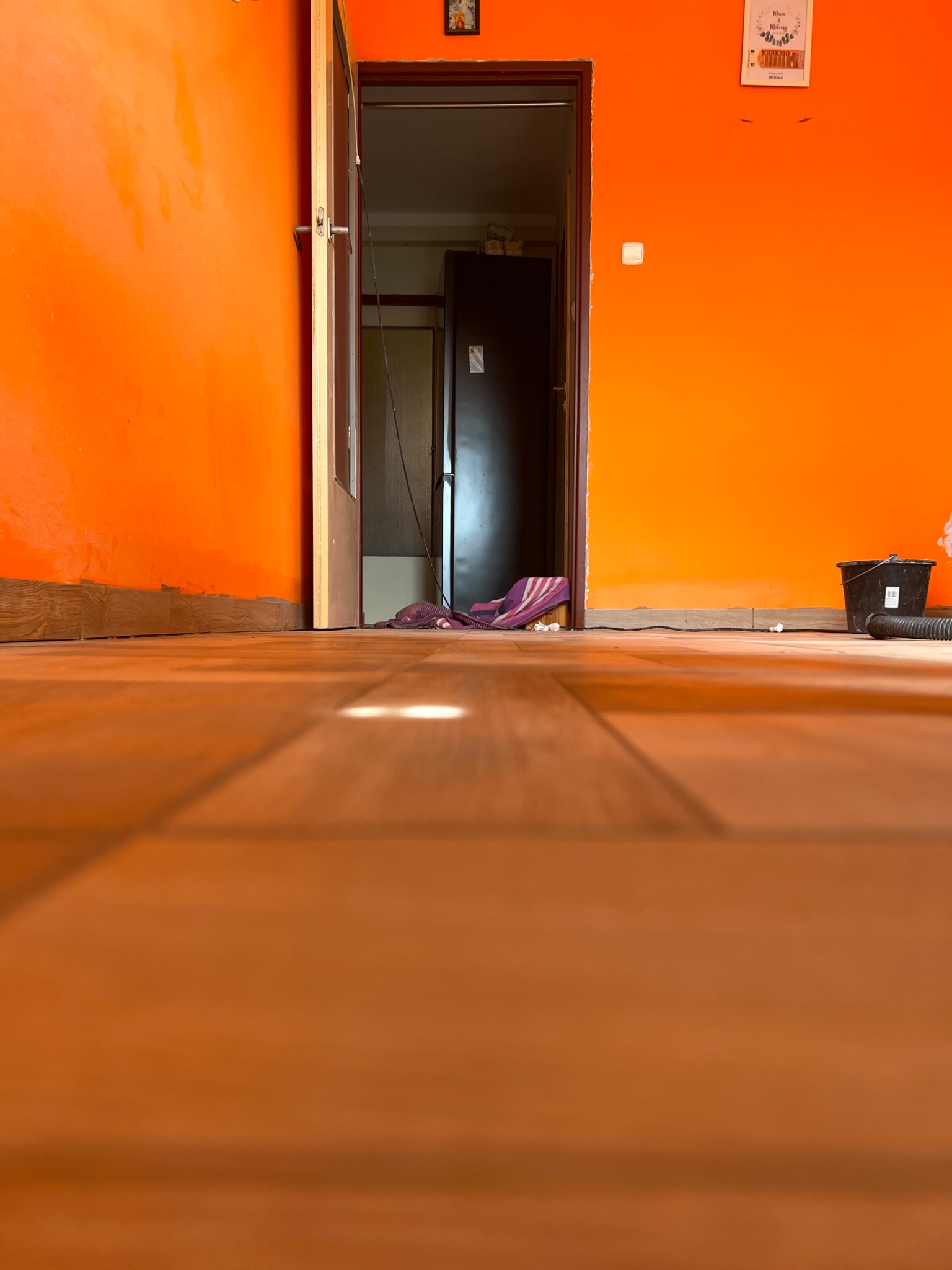 Pomarańczowe ściany i drewniana podłoga z widocznym wejściem do innego pokoju, gdzie widać ciemne drzwi i wiadro z wężem do odkurzacza.