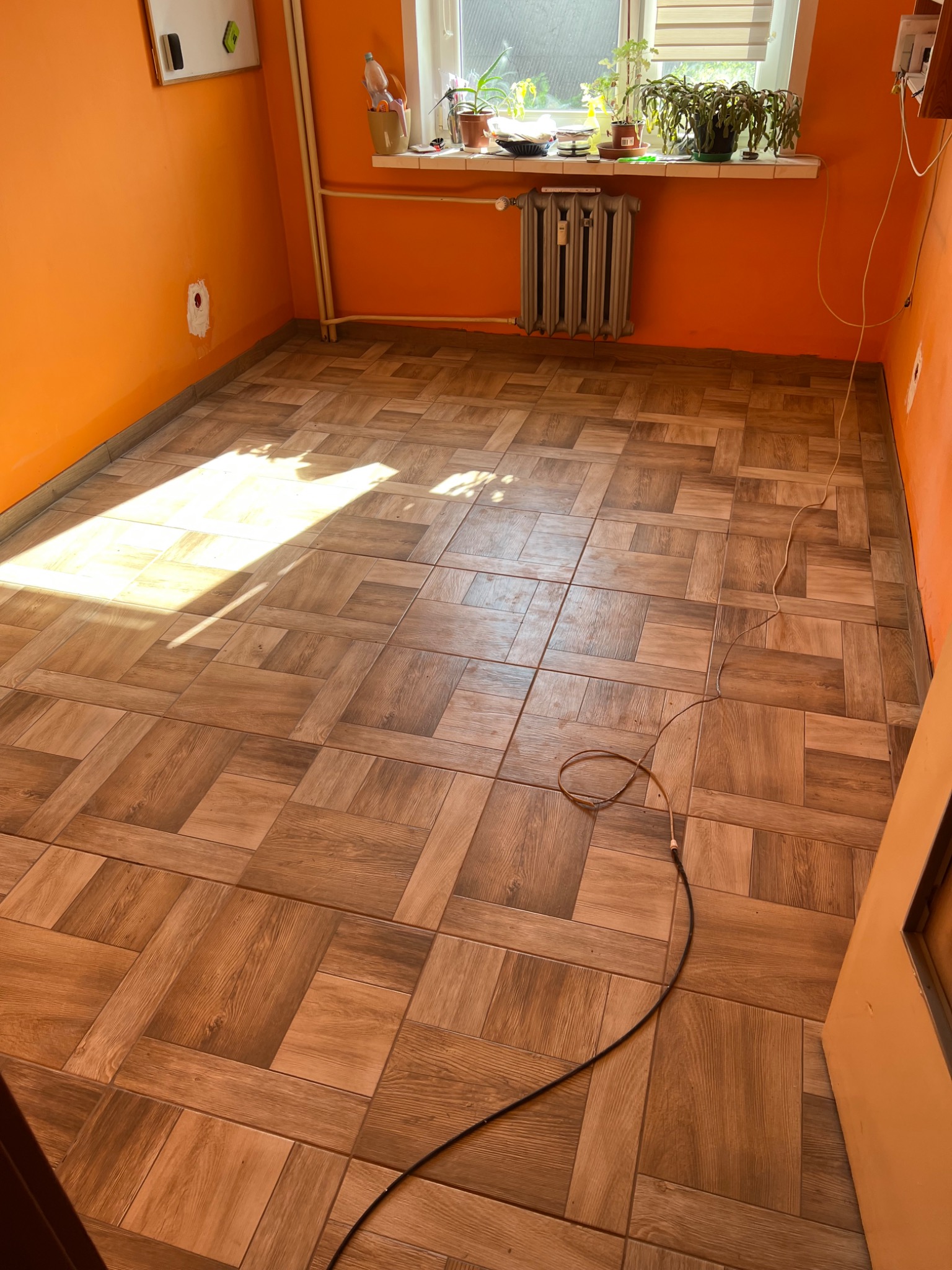 Ułożona podłoga z płytek imitujących drewno w pomarańczowym pokoju z grzejnikiem pod oknem, widoczny kabel na podłodze.