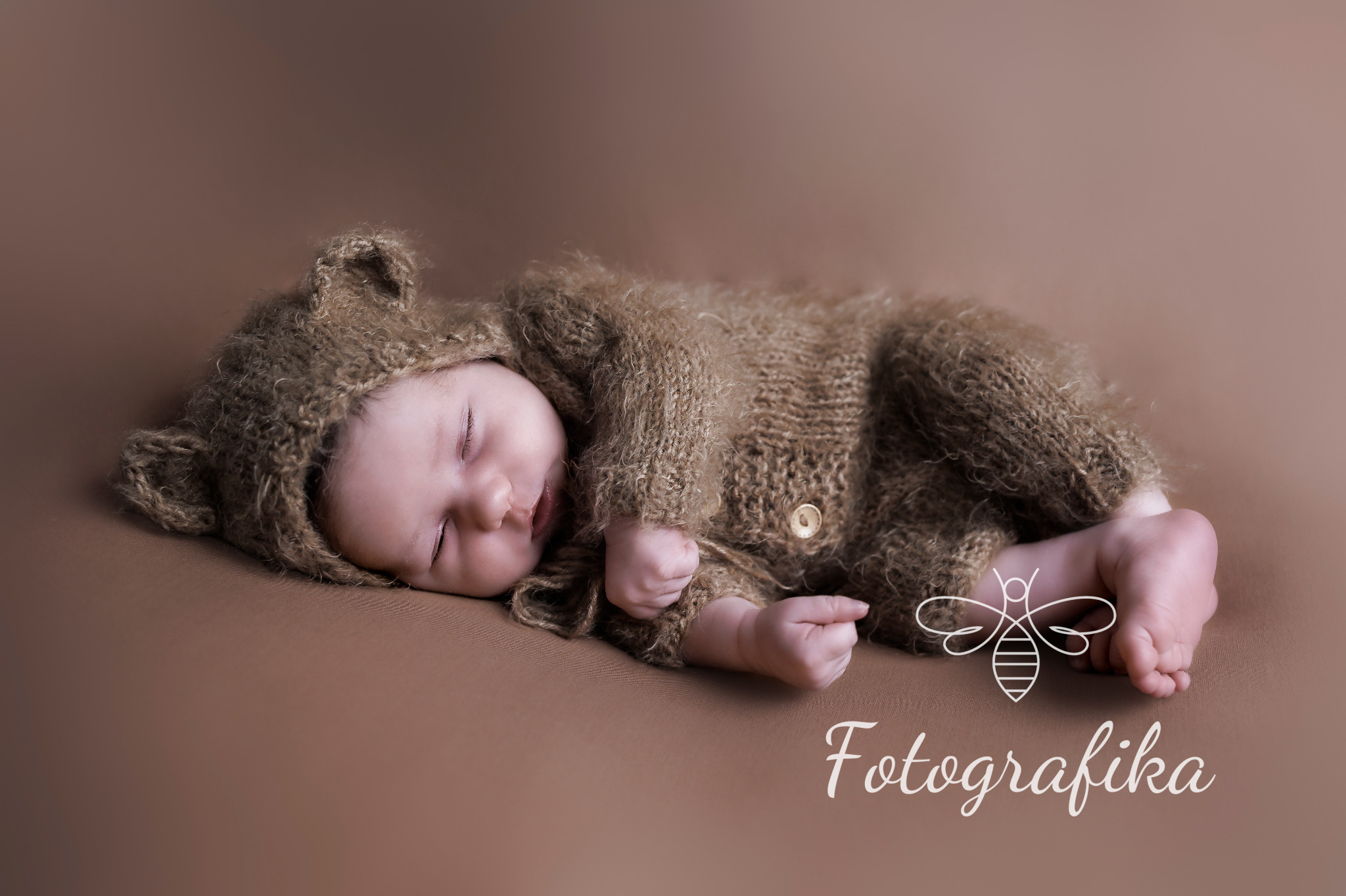 Śpiące nowonarodzone dziecko ubrane w brązowy, dziergany kombinezon z kapturem w kształcie uszu misia, leżące na brązowym tle. W prawym dolnym rogu widoczne logo fotografa.