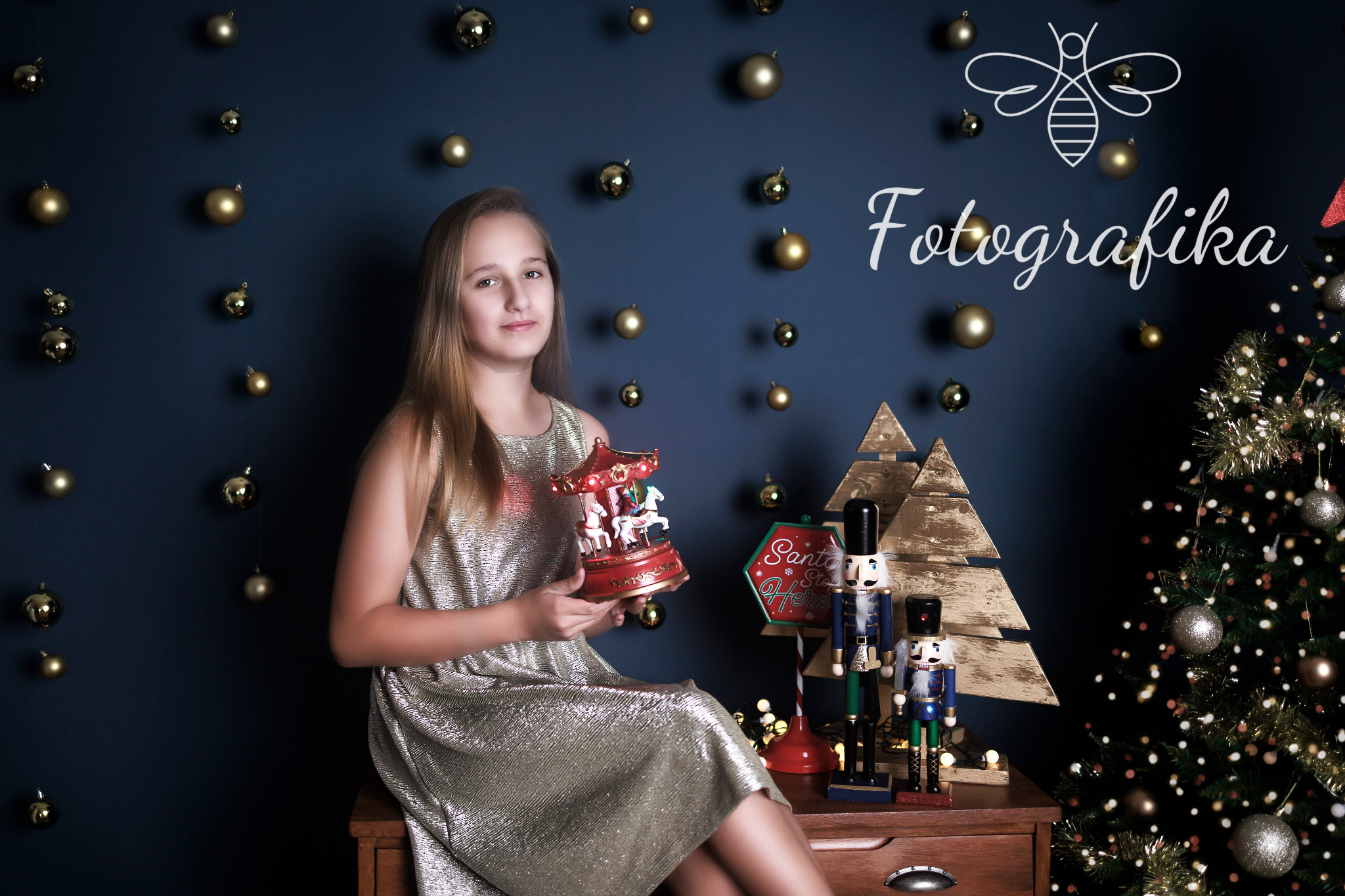 Portret dziewczynki w złotej sukience trzymającej podświetloną karuzelę z konikami na tle świątecznej dekoracji z choinką, bombkami i dziadkami do orzechów.
