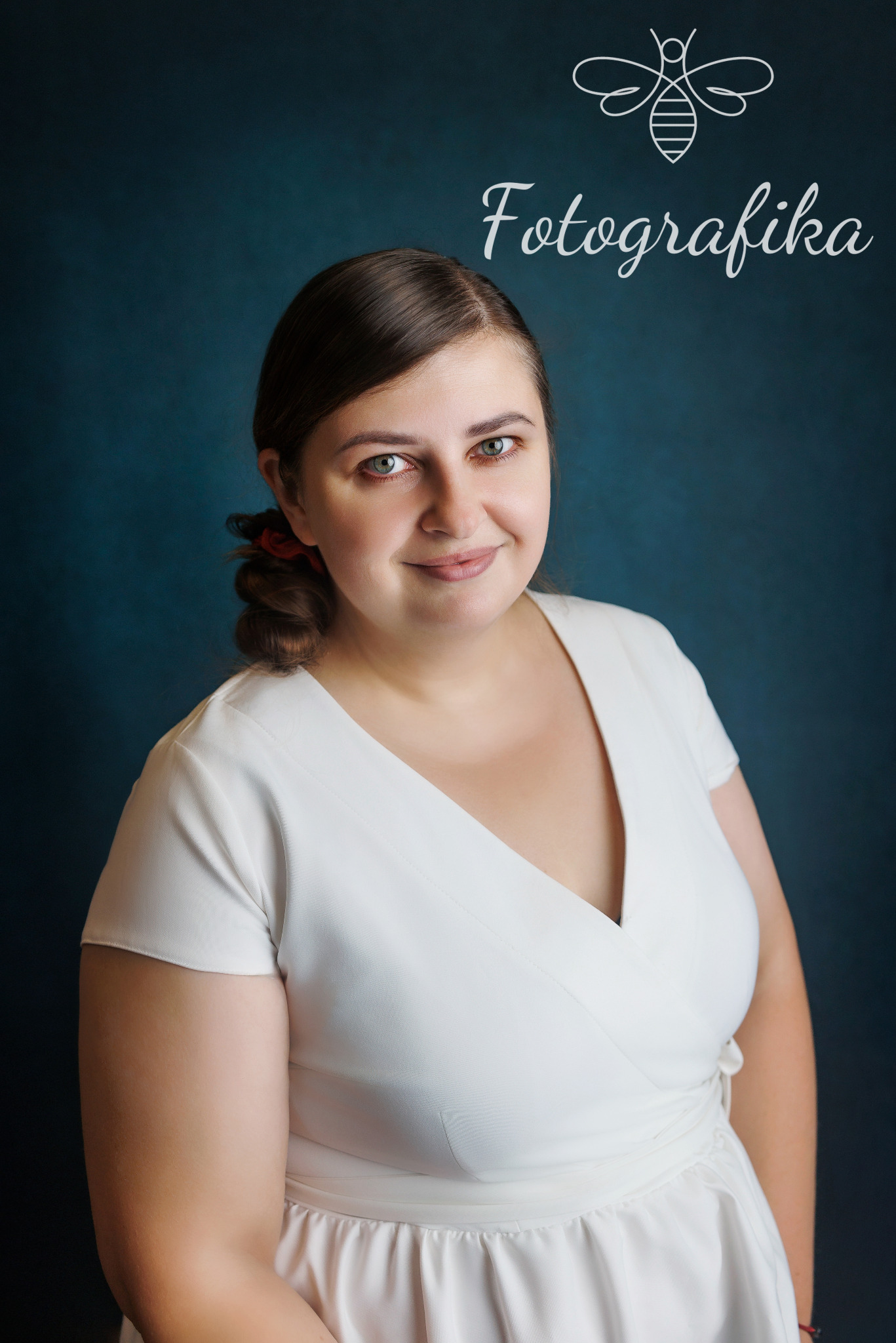 Portret uśmiechniętej kobiety w białej sukience na ciemnym tle, z logo firmy fotograficznej w górnej części kadru.
