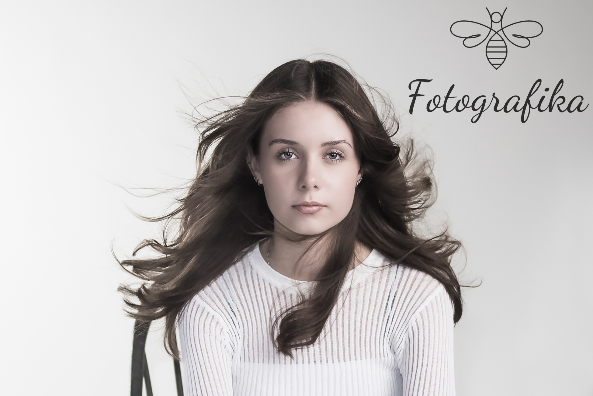 Portret młodej kobiety z rozwianymi włosami na jasnym tle, z widocznym logo firmy fotograficznej w prawym górnym rogu.