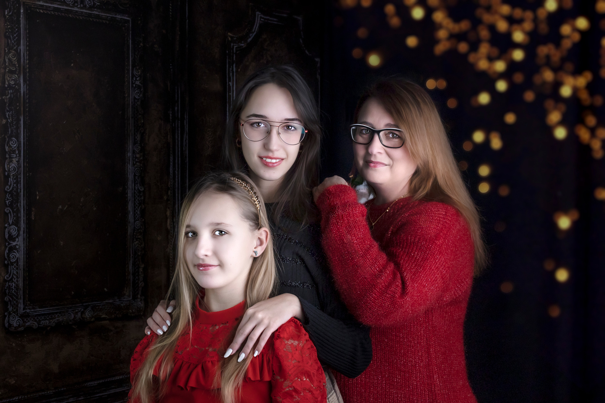 Portret rodzinny trzech kobiet w studio na tle ciemnej ściany z dekoracyjnymi lampkami bokeh, najmłodsza w czerwonej sukience z koronki, nastolatka w czarnej bluzce i okularach, matka w czerwonym...