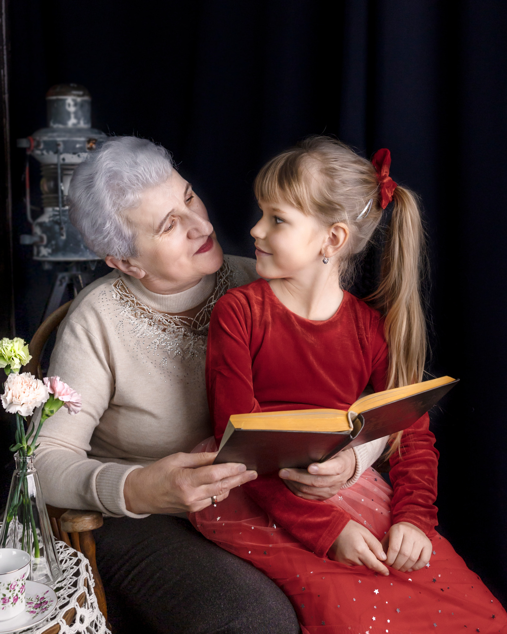 Babcia z siwymi włosami czyta wnuczce w czerwonej sukience książkę, patrząc na nią z miłością, w tle starodawna lampa studyjna