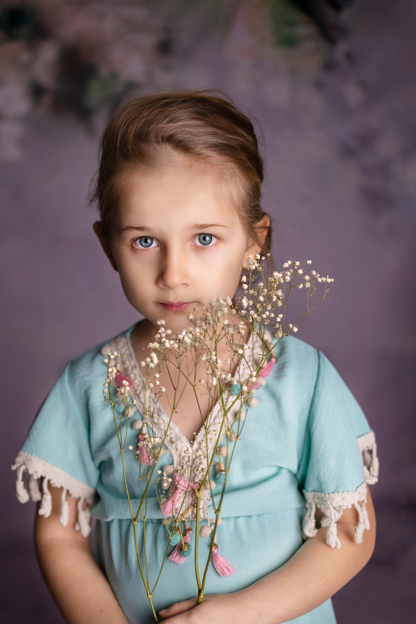 Portret dziewczynki o niebieskich oczach, trzymającej bukiecik gipsówki przed sobą, w błękitnej sukience z koronkowymi detalami i różowymi pomponami na tle fioletowego tła.