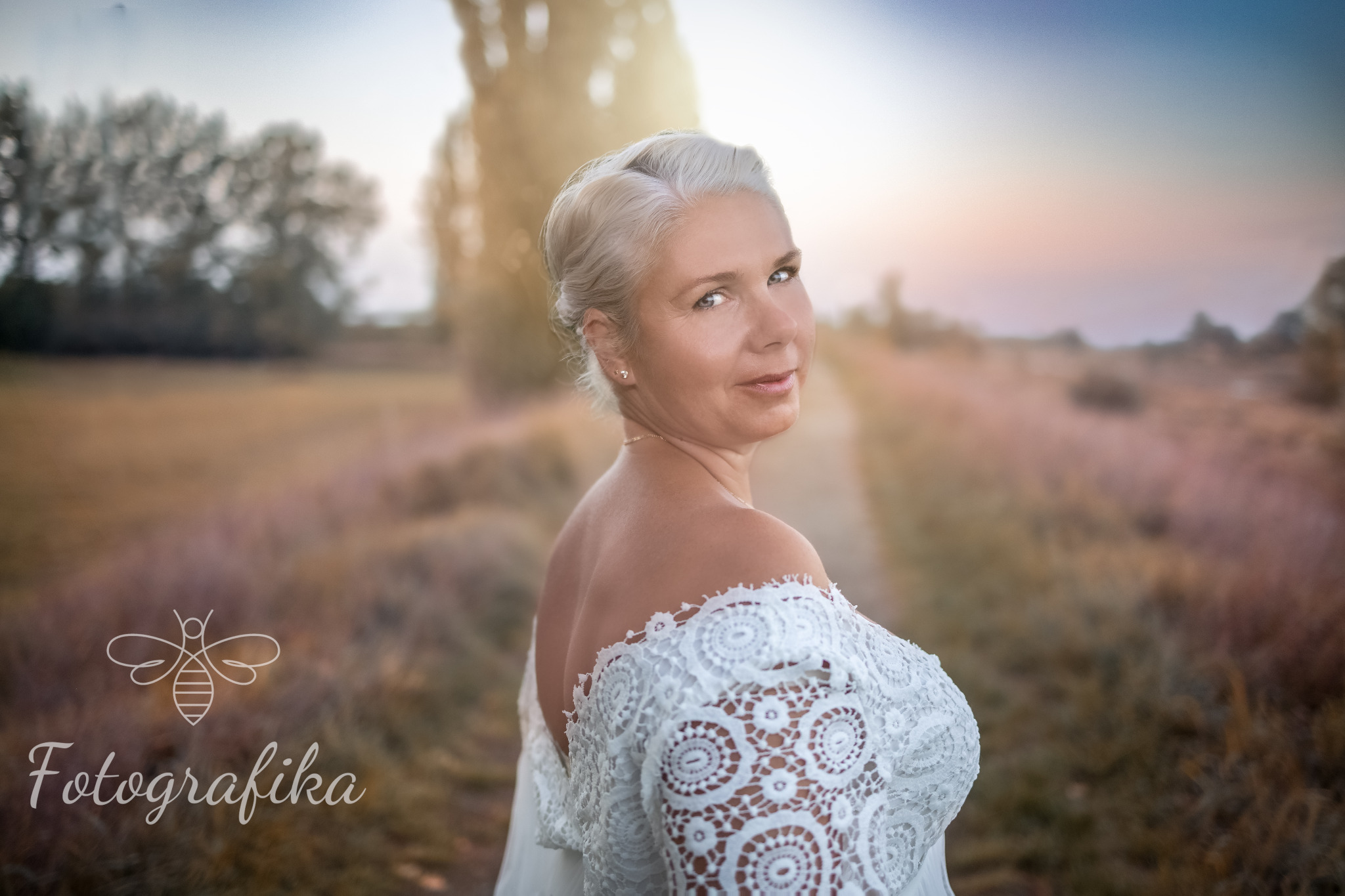 Portret kobiety w białej koronkowej sukience, spoglądającej przez ramię na tle polnej drogi o zachodzie słońca w Oławie.