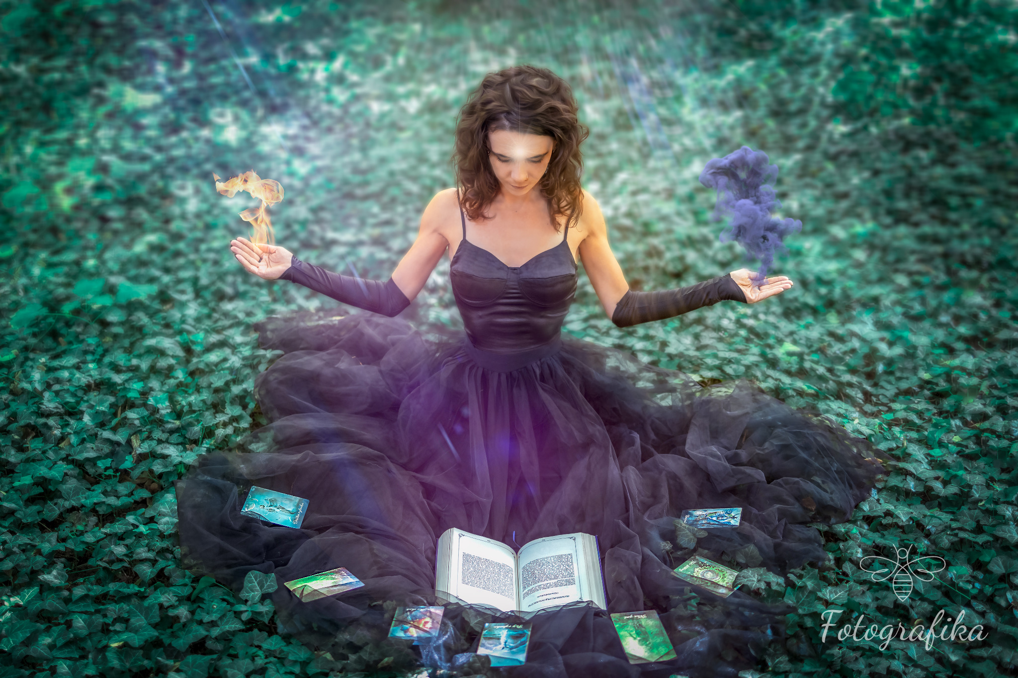 Kobieta w czarnej sukni i rękawiczkach siedzi na tle bluszczu z otwartą księgą i kartami tarota, unosząc dłonie z efektami specjalnymi ognia i dymu.