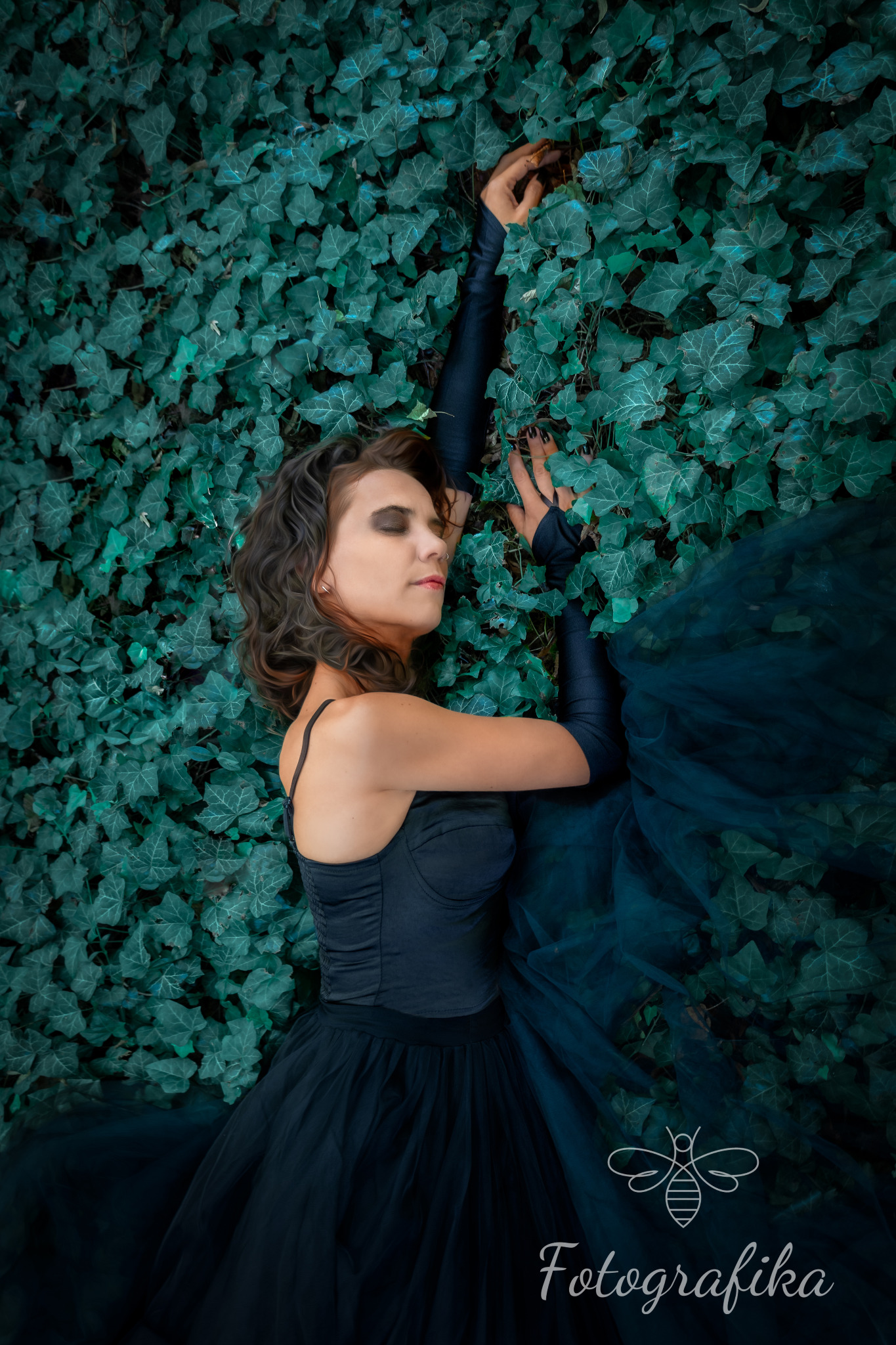 Kobieta w czarnej sukni leżąca na tle ściany z bluszczu, z rękami uniesionymi do góry i zamkniętymi oczami, stylizowana sesja zdjęciowa.