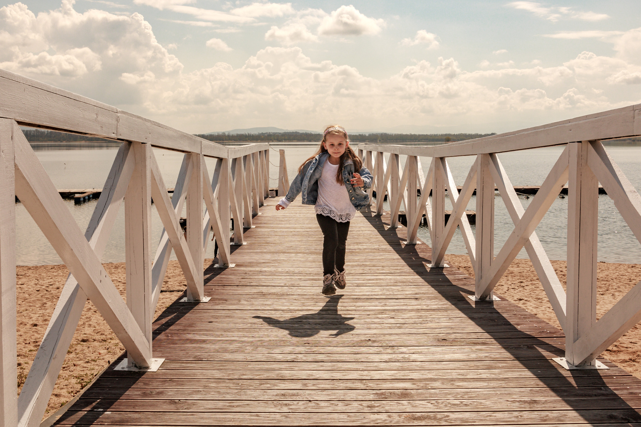 Dziewczynka biegnie po drewnianym molo w kierunku obiektywu, z widokiem na jezioro i chmurne niebo w tle.