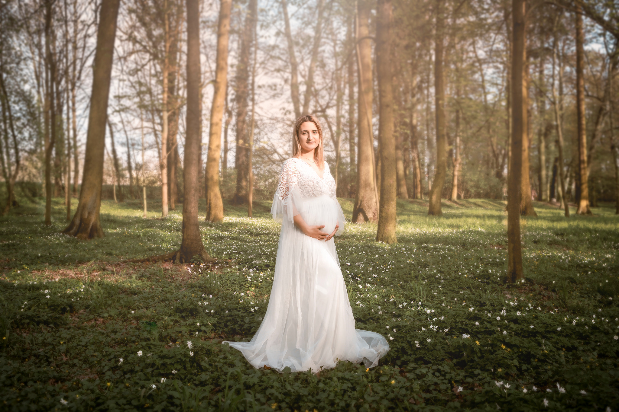 Kobieta w białej sukni ciążowej pozuje w lesie pełnym kwitnących białych kwiatów, trzymając dłonie na brzuchu.