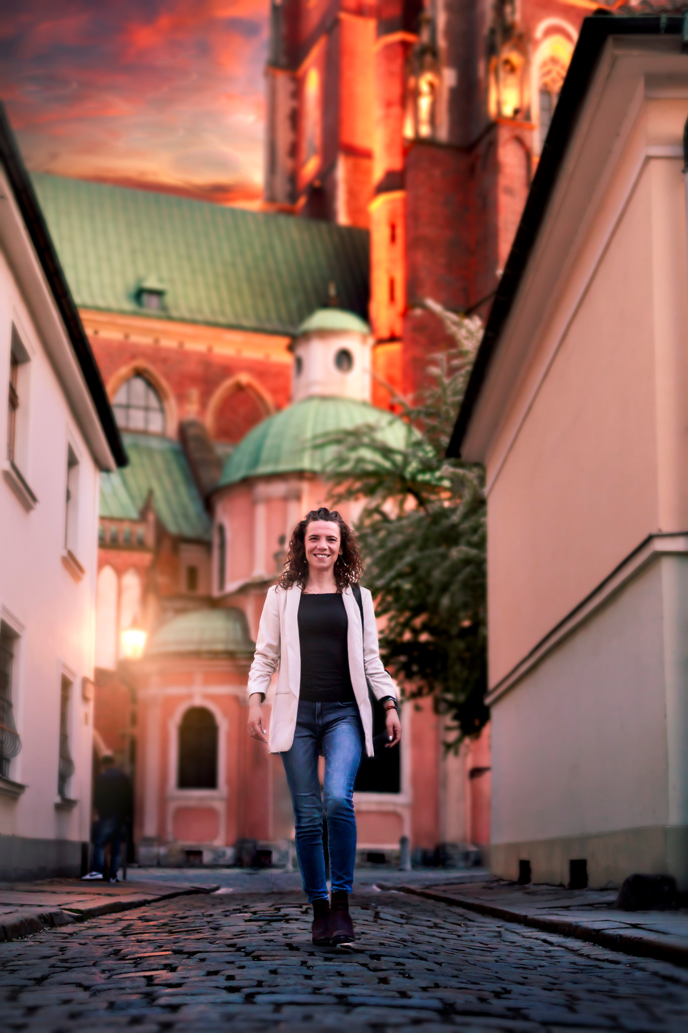 Uśmiechnięta kobieta w jeansach i jasnej marynarce spaceruje brukowaną uliczką, w tle widoczny zabytkowy kościół z zielonymi kopułami i czerwonymi wieżami.