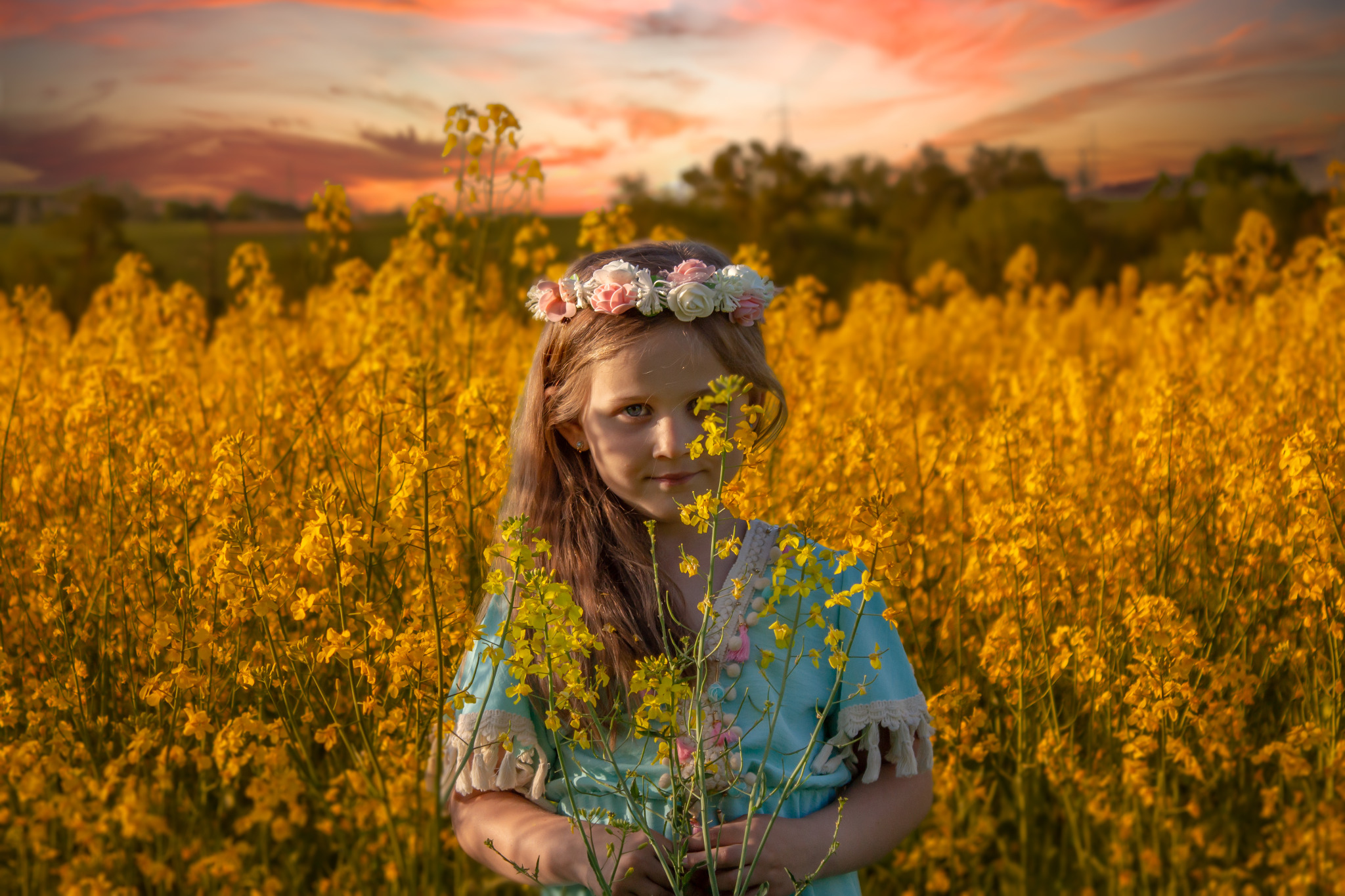 Dziewczynka z wiankiem na głowie, ubrana w błękitną sukienkę, stoi na tle pola rzepaku podczas zachodu słońca, trzymając w dłoniach kwitnącą gałązkę rzepaku.