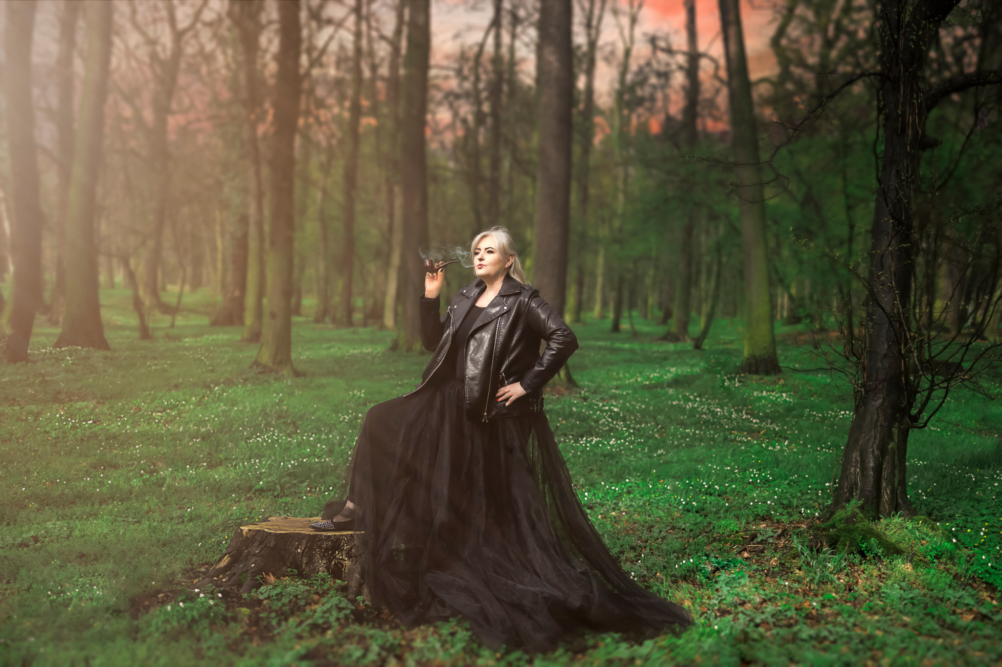 Kobieta z siwymi włosami w czarnej skórzanej kurtce i tiulowej spódnicy siedzi na pniu w lesie, paląc fajkę.
