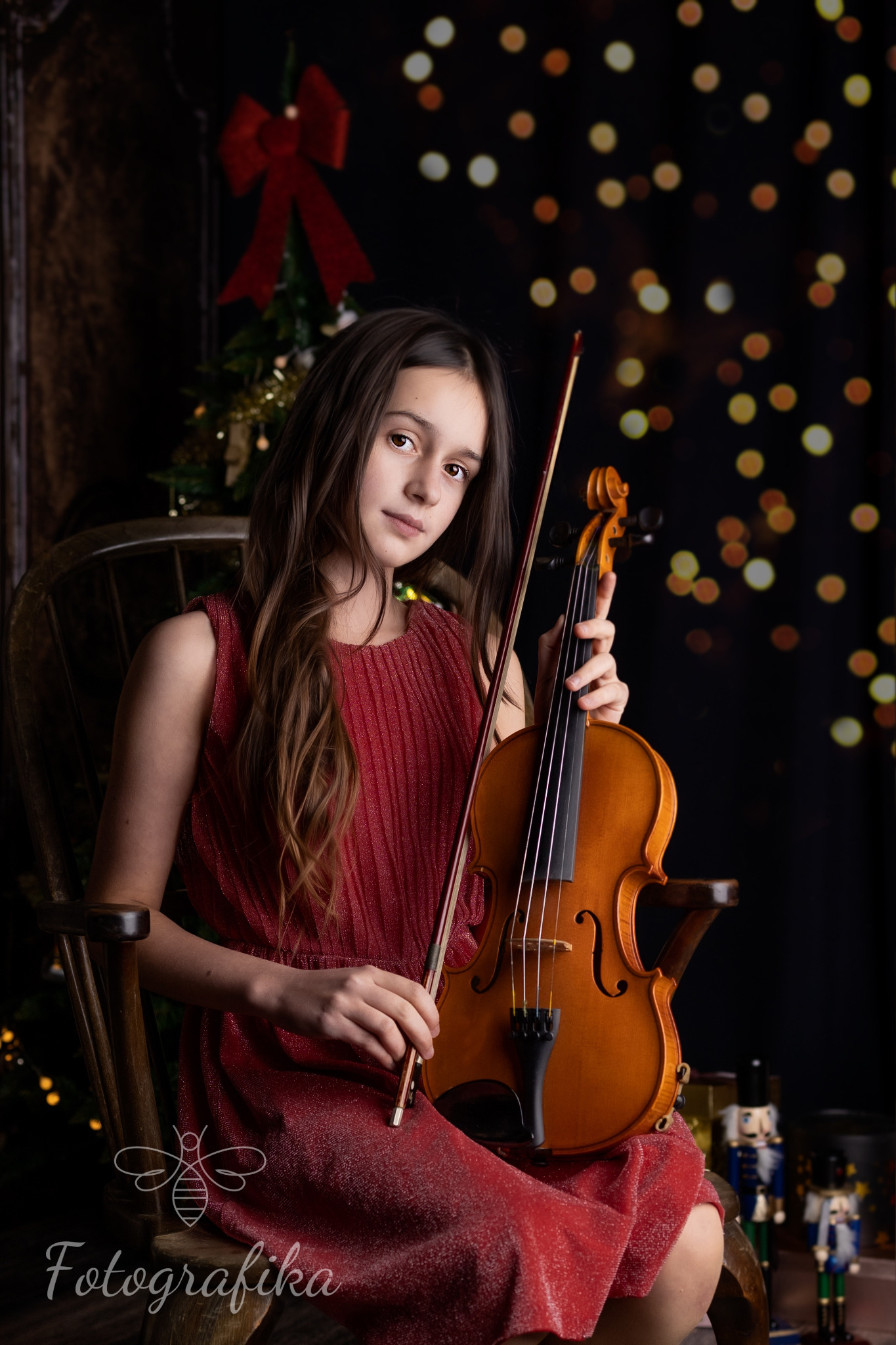 Młoda dziewczyna w czerwonej sukience siedzi na drewnianym krześle, trzymając skrzypce na tle świątecznej scenerii z choinką i światełkami bokeh.