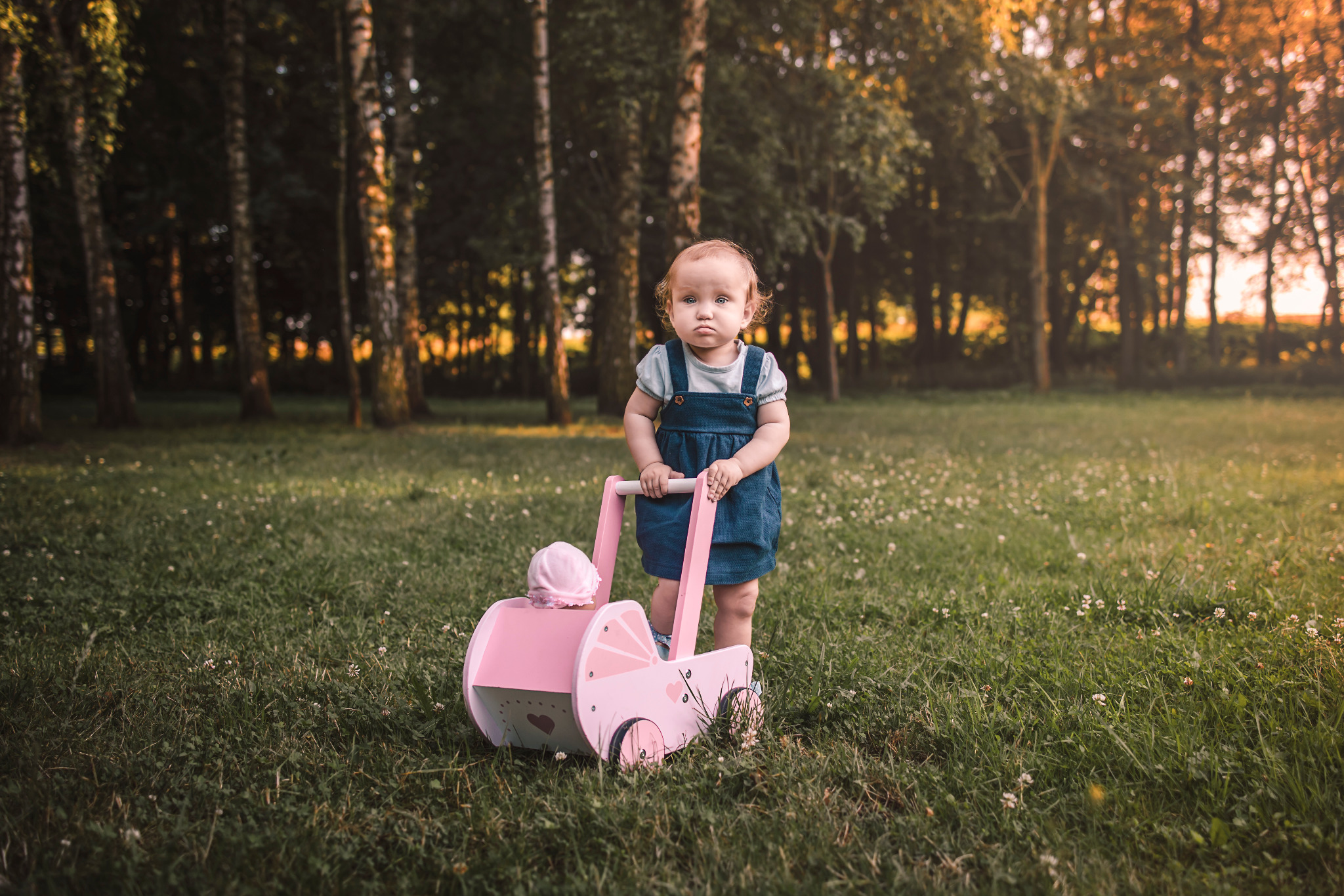 Mała dziewczynka w dżinsowej sukience pcha różowy, drewniany wózek dla lalek na trawiastym terenie w otoczeniu drzew w promieniach zachodzącego słońca.