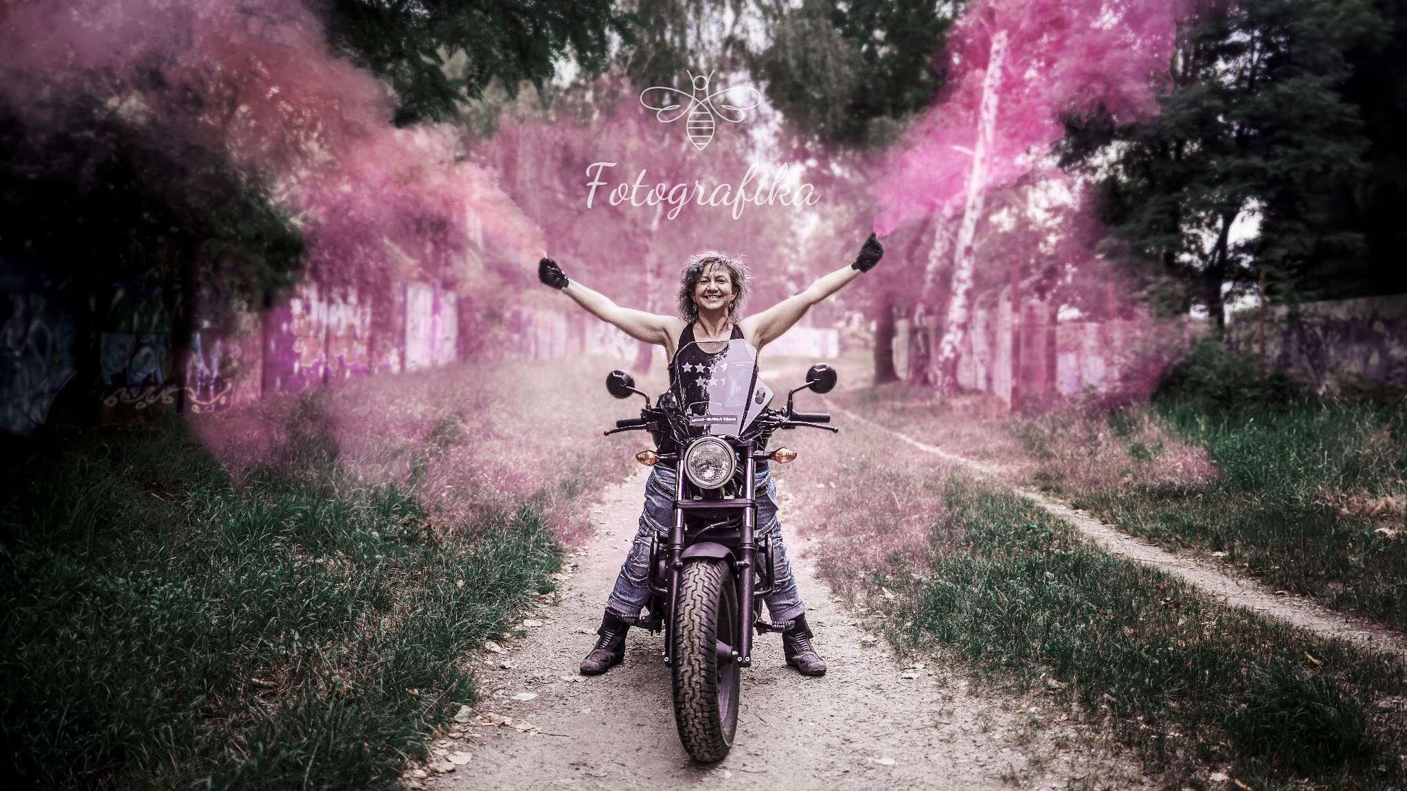 Kobieta z rozpostartymi ramionami siedzi na motocyklu, trzymając w dłoniach dymy koloru różowego na polnej drodze.