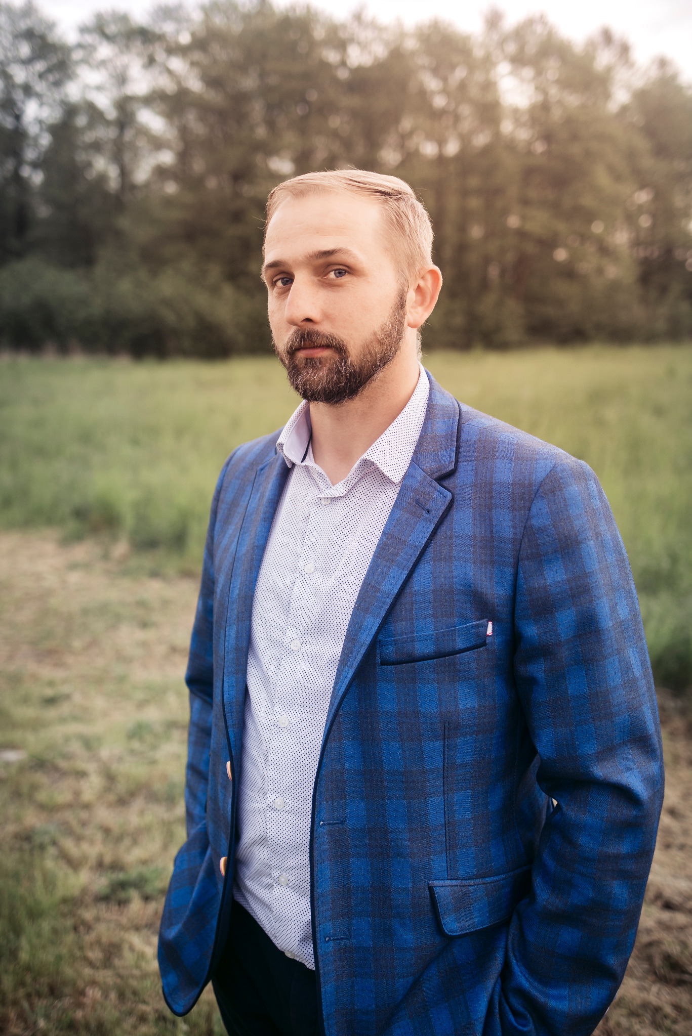 Mężczyzna z brodą w niebieskiej marynarce w kratę i koszuli w kropki, stojący na łące z drzewami w tle, patrzy w obiektyw.