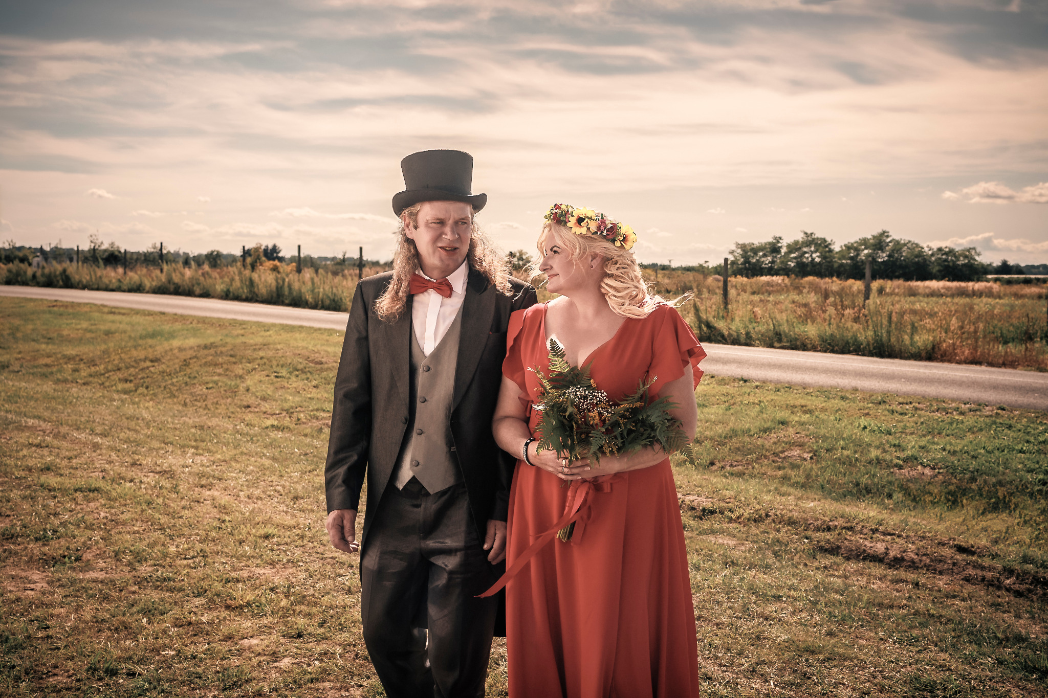 Para młoda idzie polną drogą: mężczyzna w cylindrze i czerwonej muszce, kobieta w czerwonej sukni z wiankiem na głowie i bukietem polnych kwiatów.