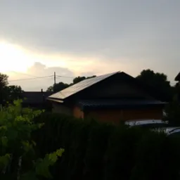 Dom jednorodzinny z panelami słonecznymi na dachu, widok zza żywopłotu i winorośli o zmierzchu.