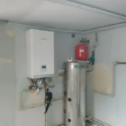 Biały piec Kaisai zawieszony na ścianie obok srebrnego zbiornika HEAT ECO z czerwonym naczyniem wzbiorczym, instalacja w surowym pomieszczeniu z widocznymi rurami.