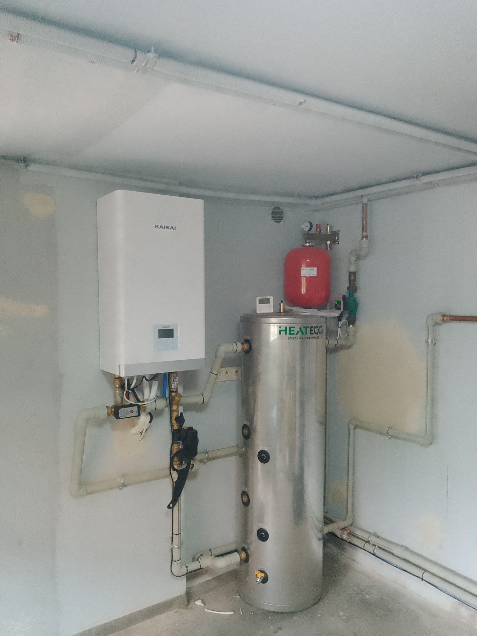 Biały piec Kaisai zawieszony na ścianie obok srebrnego zbiornika HEAT ECO z czerwonym naczyniem wzbiorczym, instalacja w surowym pomieszczeniu z widocznymi rurami.