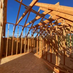 Wnętrze domu szkieletowego w trakcie budowy, widoczne drewniane belki konstrukcyjne na tle błękitnego nieba. Podłoga z płyt OSB, fragmenty drewna w dolnym rogu.