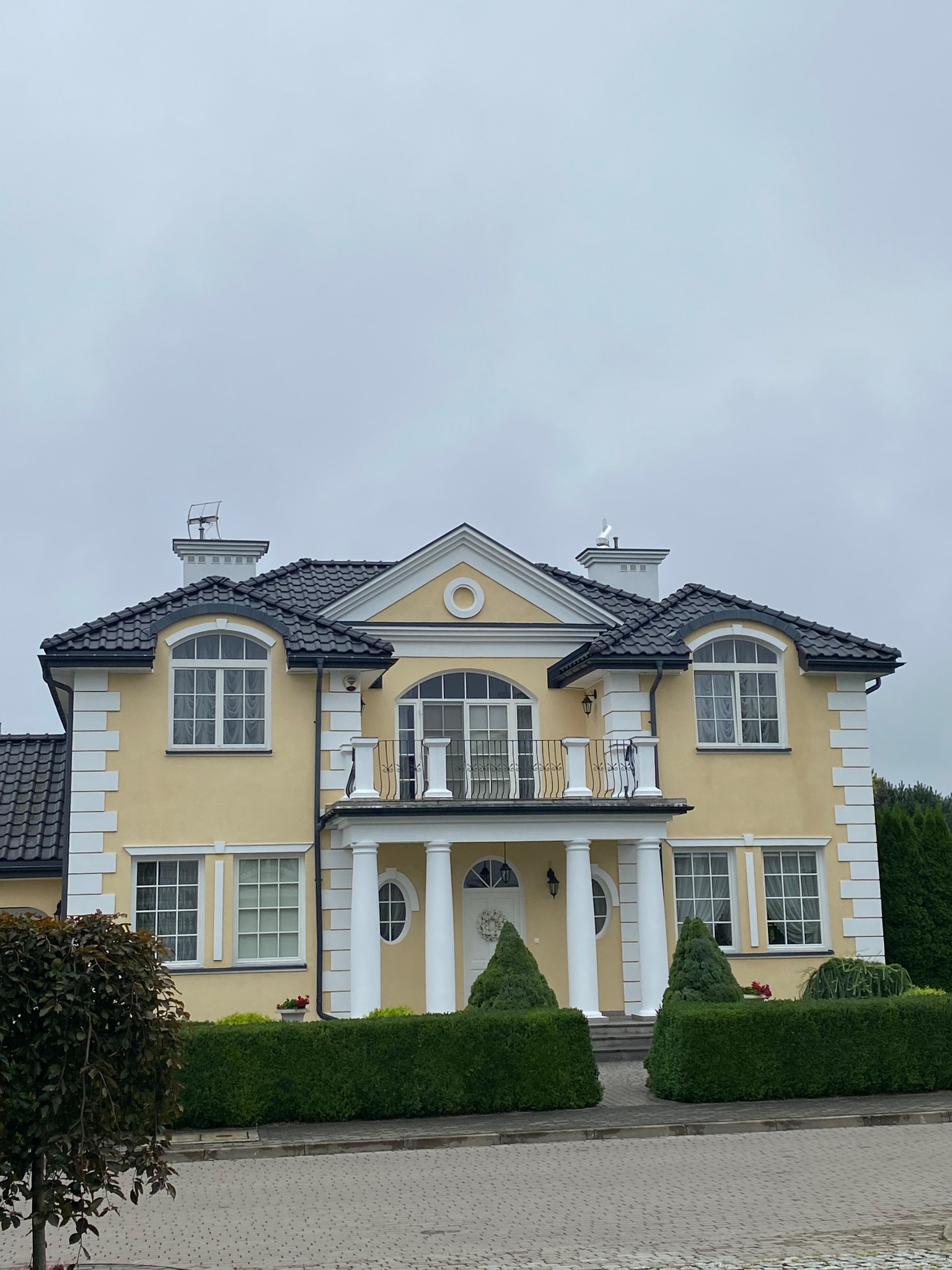 Żółta elewacja dwukondygnacyjnego domu z ciemną dachówką, białymi kolumnami i balkonem z czarną balustradą, otoczona zielonymi krzewami i drzewami.