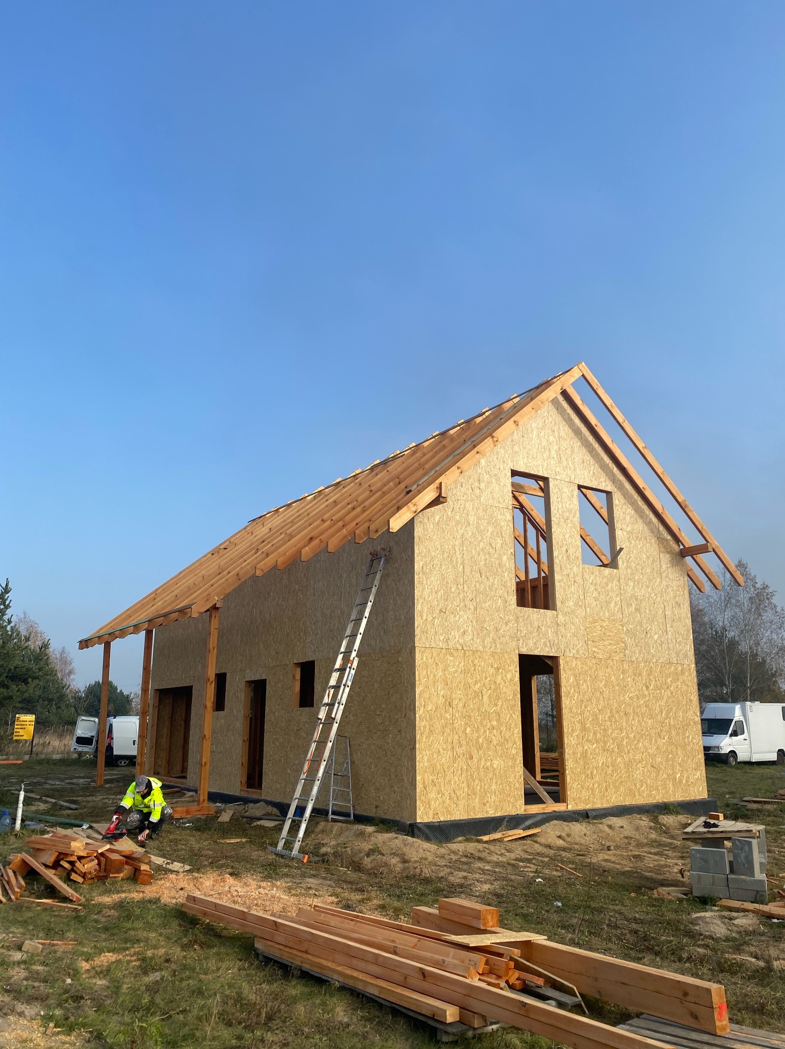 Budowa domu szkieletowego w trakcie realizacji: widoczne ściany z płyt OSB, drewniana konstrukcja dachu i pracownik w odblaskowej kurtce przycinający drewno.