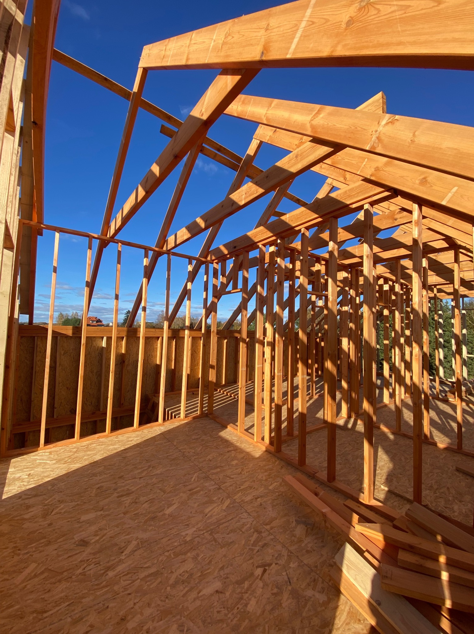Wnętrze domu szkieletowego w trakcie budowy, widoczne drewniane belki konstrukcyjne na tle błękitnego nieba. Podłoga z płyt OSB, fragmenty drewna w dolnym rogu.