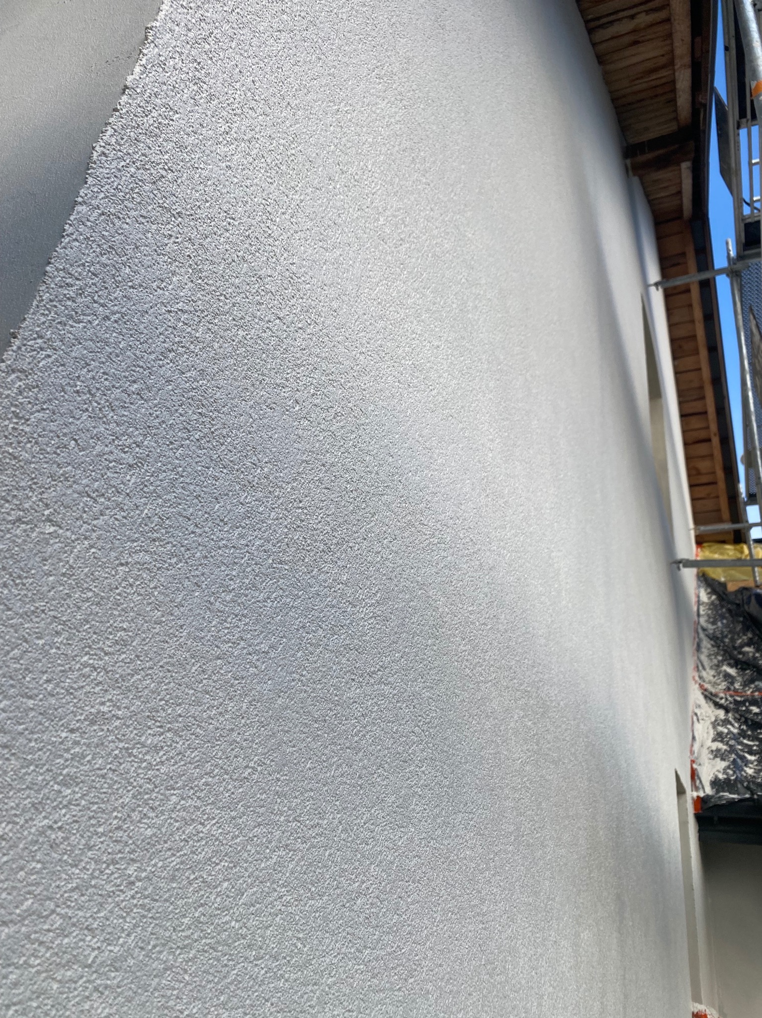 Świeżo nałożony, jasnoszary tynk strukturalny na elewacji domu, widoczny z bliska pod kątem. Fragment rusztowania i drewnianej konstrukcji dachu w tle.