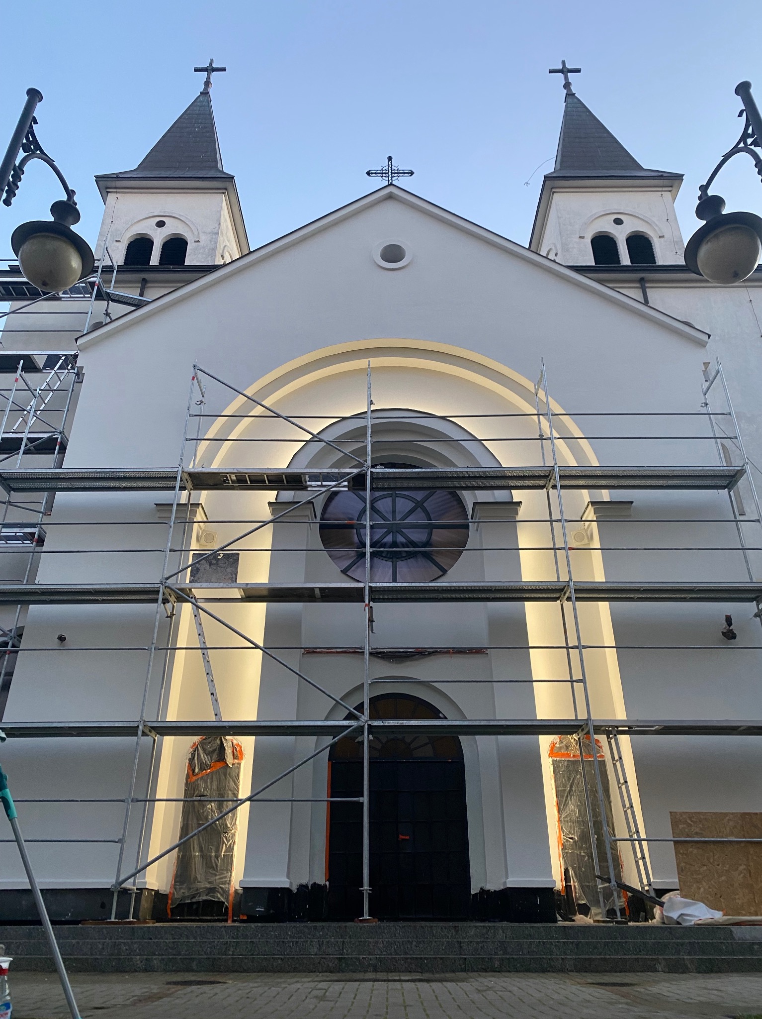 Biała fasada kościoła w trakcie renowacji, z rusztowaniami zasłaniającymi część elewacji. Widoczne wieże z krzyżami i okrągłe okno nad wejściem.