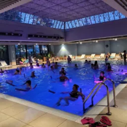 Grupowe zajęcia aqua aerobiku w basenie z niebieskim oświetleniem, uczestnicy używają piankowych makaronów, w tle leżaki i osoby odpoczywające.