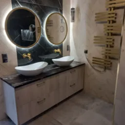 Nowoczesna łazienka z dwoma okrągłymi lustrami podświetlanymi LED, umywalkami nablatowymi w kształcie łódki, blatem z czarnego marmuru i grzejnikiem drabinkowym w kolorze złotym.