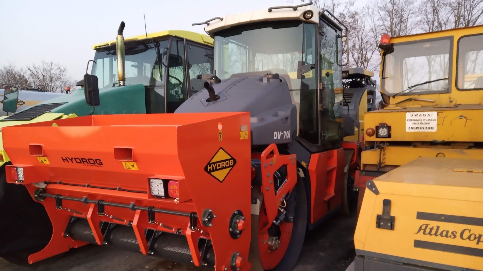 Pomarańczowa maszyna drogowa z logiem Hydrog, widoczna z tyłu, obok stoją inne maszyny budowlane w kolorach żółtym i zielonym, na tle widoczne drzewa bez liści.