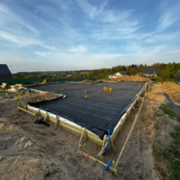 Przygotowany zbrojony szalunek pod płytę fundamentową z widocznymi prętami zbrojeniowymi i rurami instalacyjnymi, otoczony drewnianą konstrukcją i ziemią, w tle zabudowa jednorodzinna i błękitne...