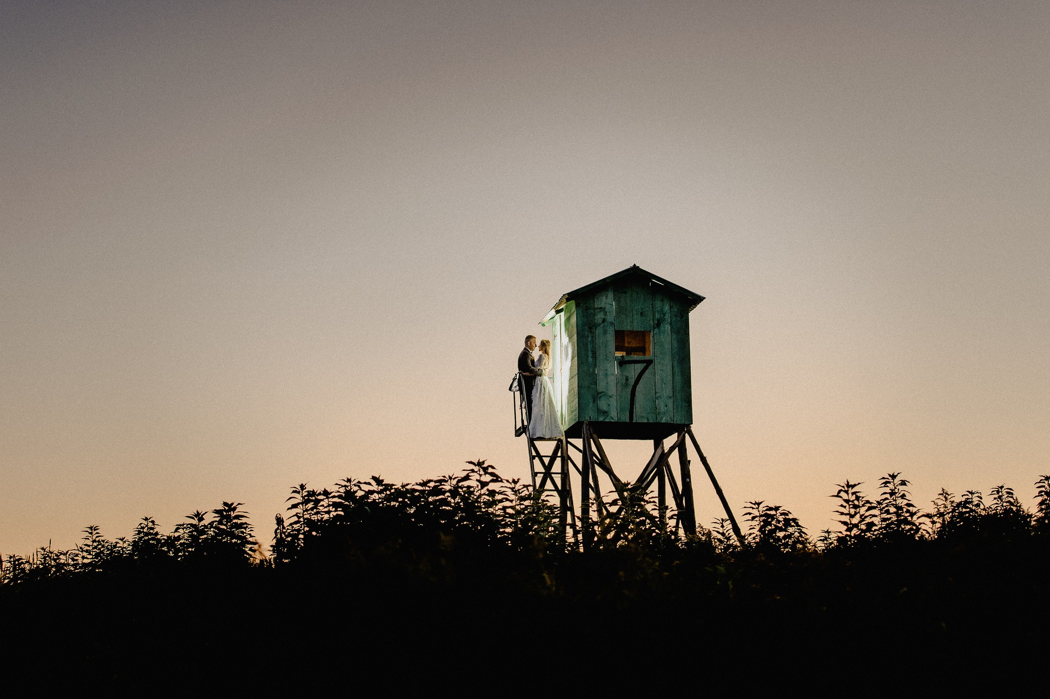 Para młoda w objęciach na platformie widokowej wśród zieleni, w tle zachodzące słońce.