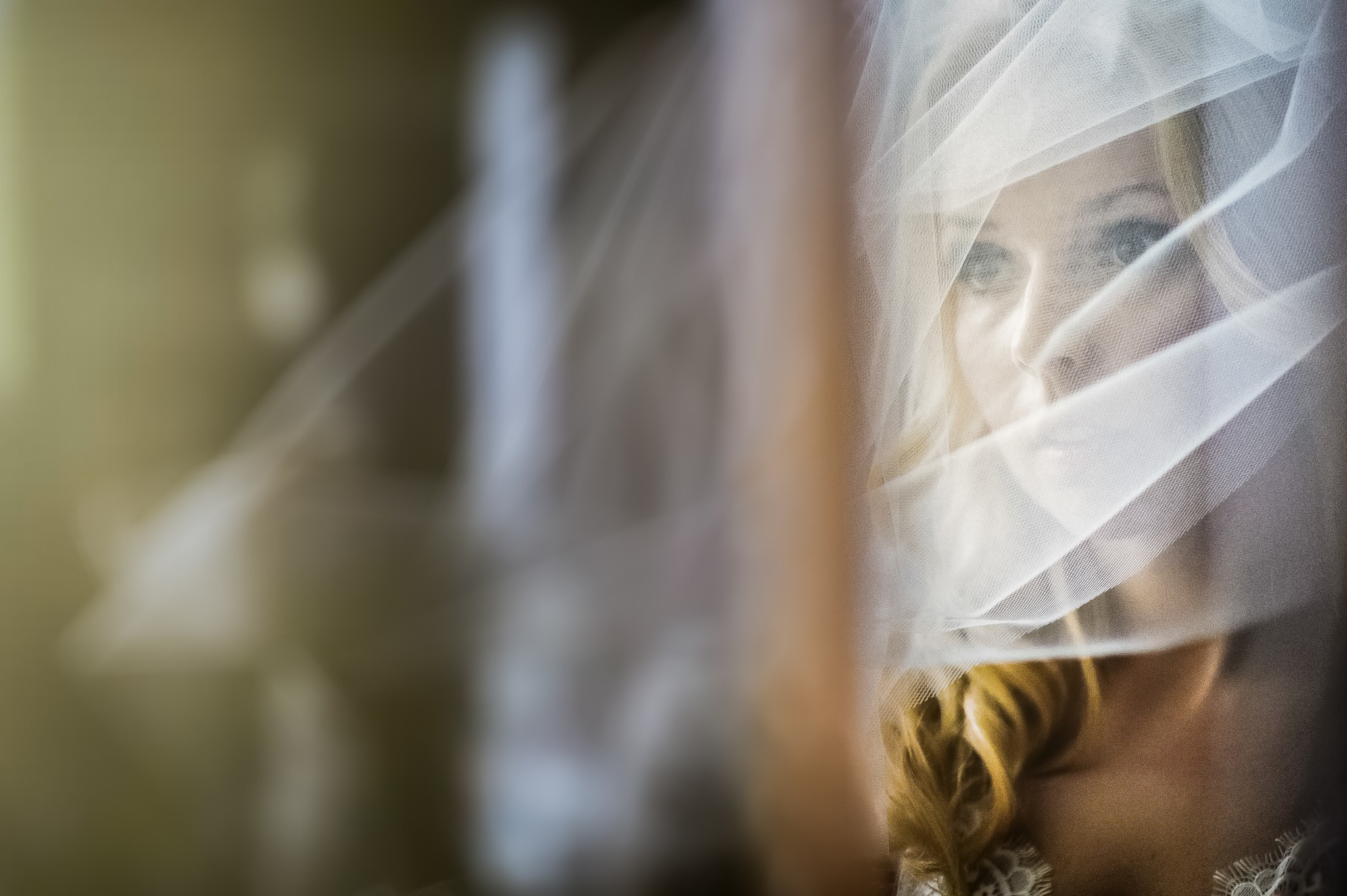 Portret panny młodej z bliska, z delikatnym welonem częściowo zasłaniającym twarz, z widocznymi niebieskimi oczami i blond włosami.