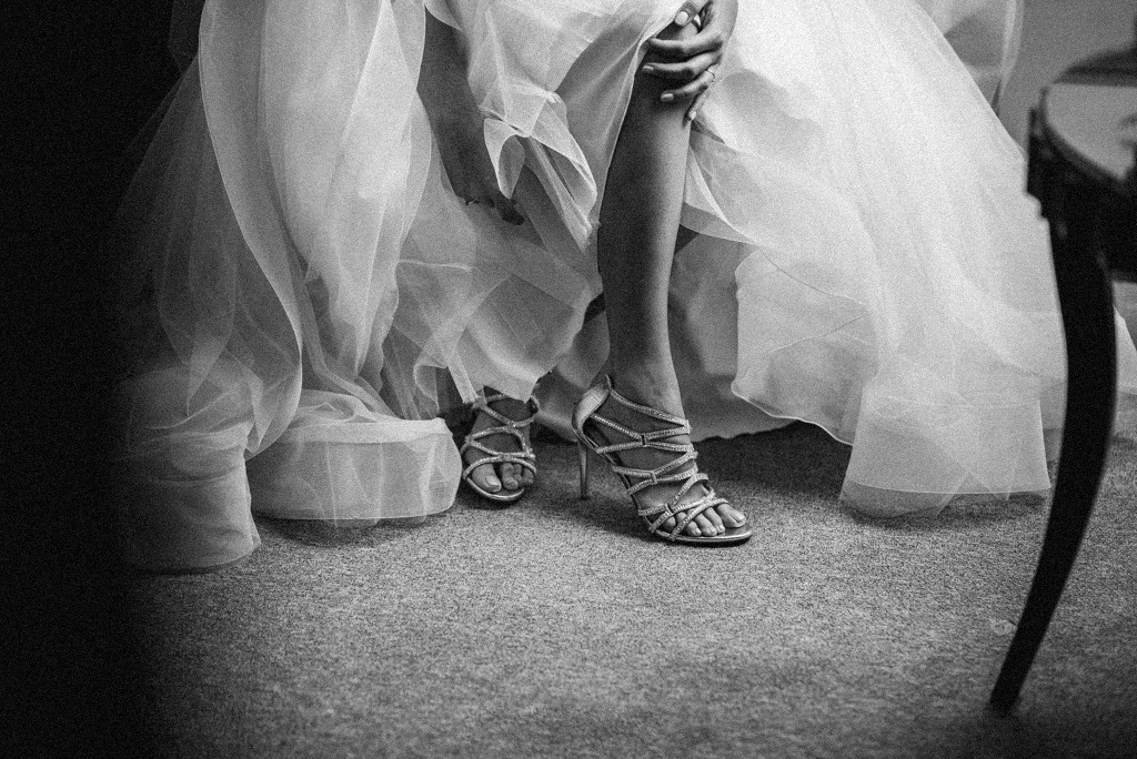 Panna młoda w sukni ślubnej i srebrnych, zdobionych sandałach na obcasie, siedząca na krześle, czarno-biała fotografia.