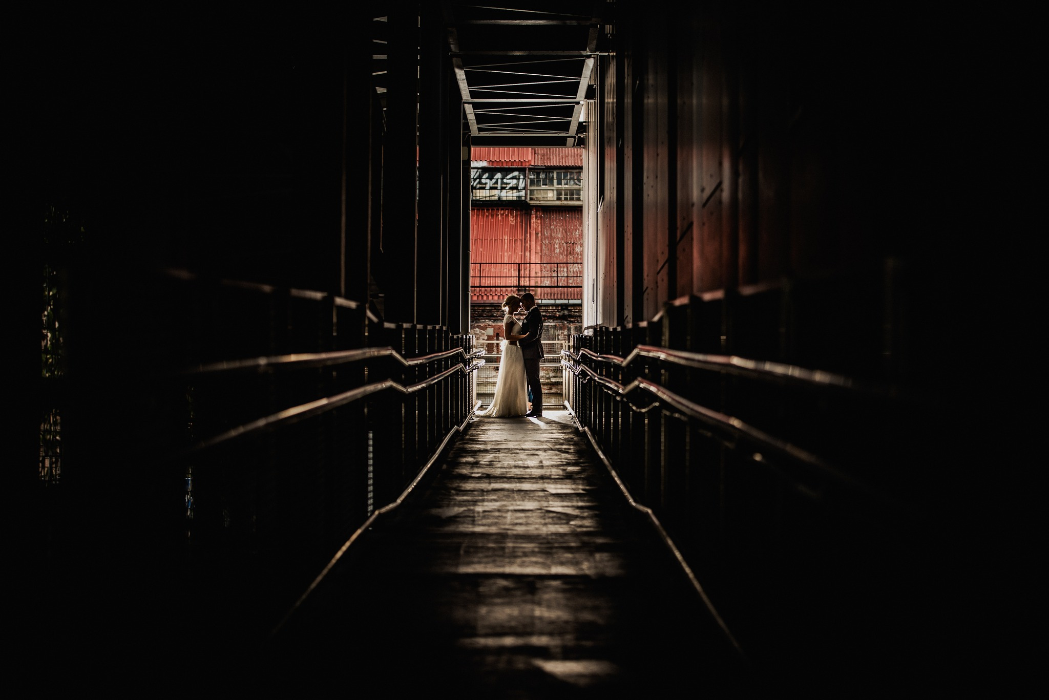 Para młoda w objęciach na tle industrialnego korytarza z metalowymi barierkami i czerwonym budynkiem w tle, ujęcie z perspektywy podłogi.