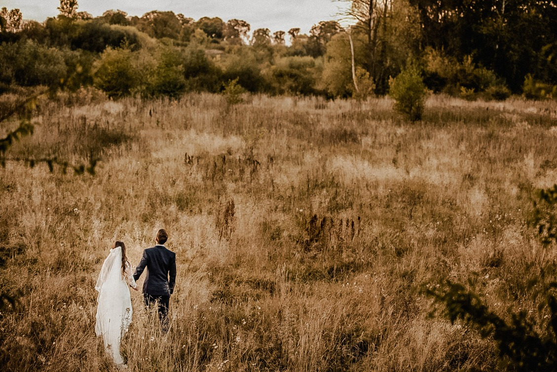 Para młoda, on w granatowym garniturze, ona w białej sukni z welonem, idąca przez wysokie trawy na tle drzew i krzewów w okolicy Ząbek, uchwyceni z tyłu.