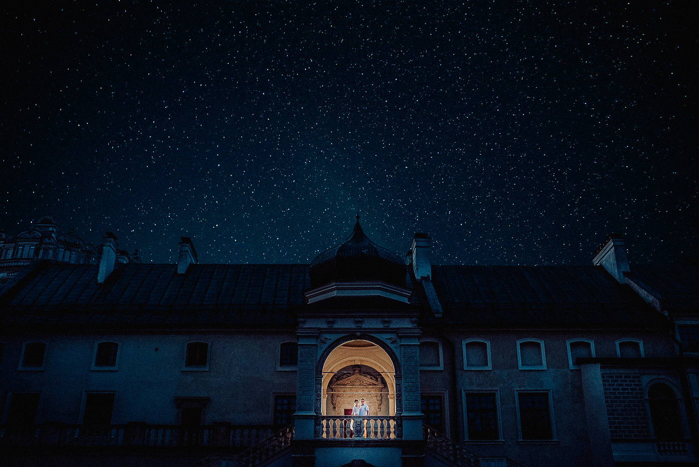 Para młoda na balkonie zamku nocą, pod rozgwieżdżonym niebem, oświetlona ciepłym światłem.