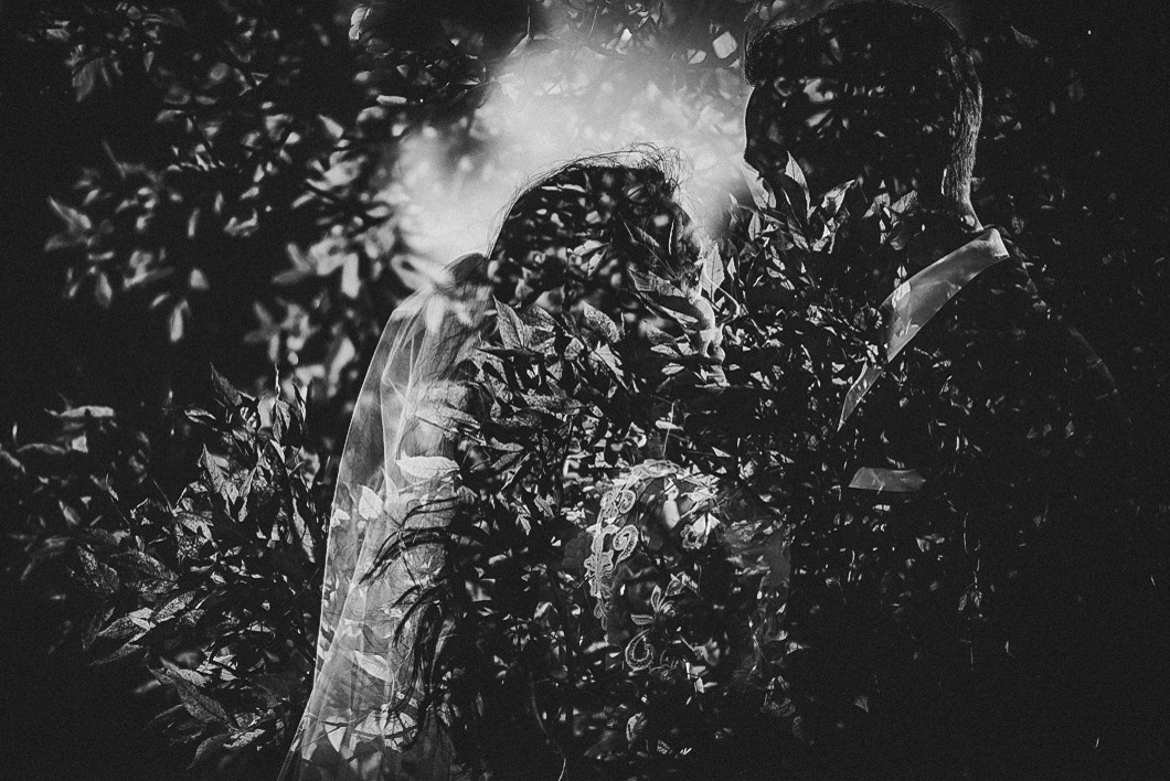 Czarno-białe, artystyczne ujęcie pary młodej ukrytej za liśćmi, z efektem nałożonych warstw, tworzące oniryczny nastrój.