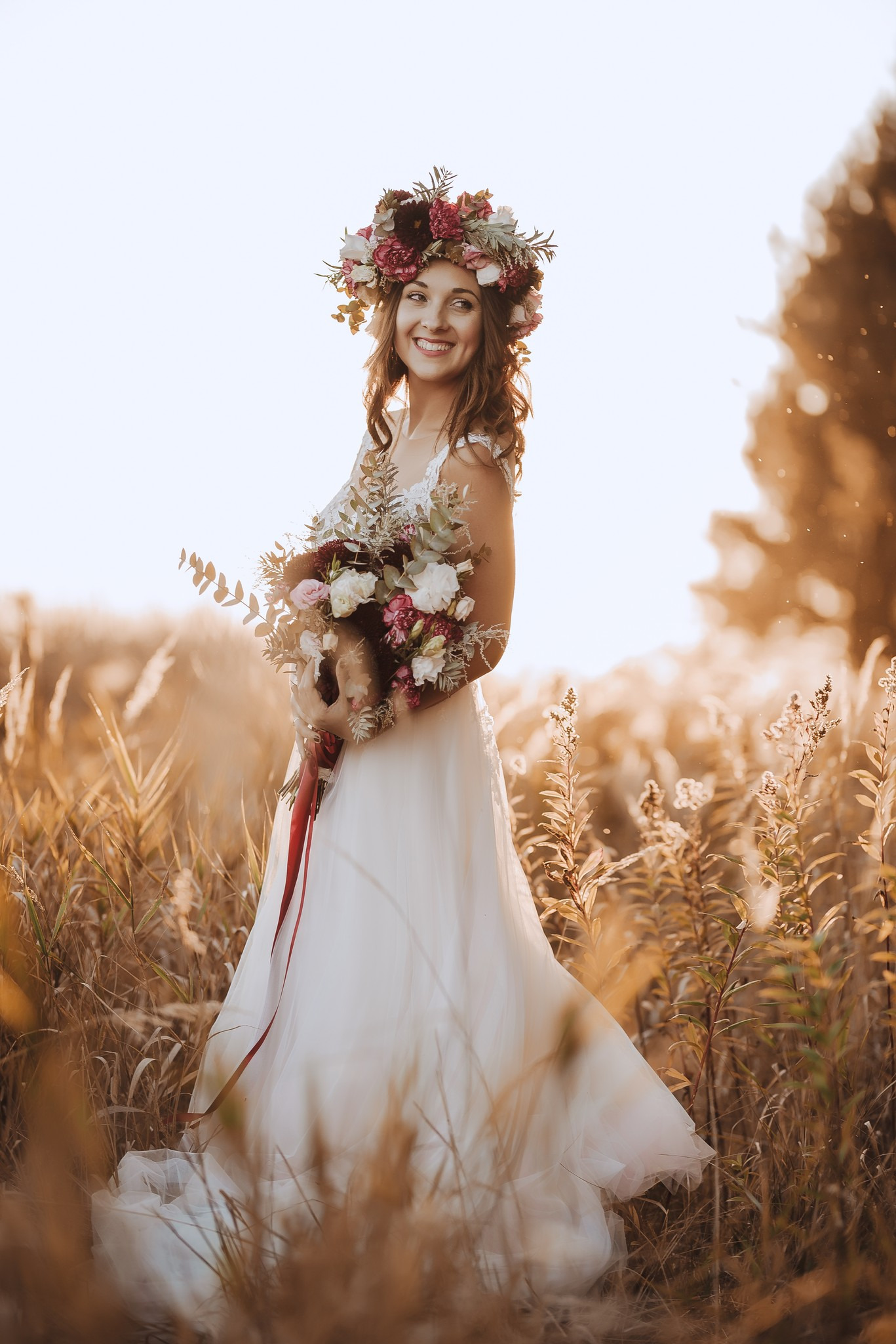 Uśmiechnięta panna młoda w białej sukni i wianku z kwiatów, trzymająca bukiet, pozuje na tle łąki w ciepłym, naturalnym świetle.