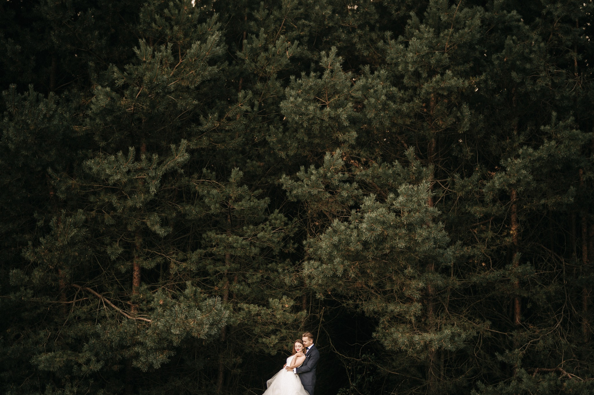 Para młoda w objęciach na tle gęstego, ciemnego lasu iglastego; panna młoda w białej sukni z odkrytymi ramionami, pan młody w ciemnym garniturze, delikatne oświetlenie.