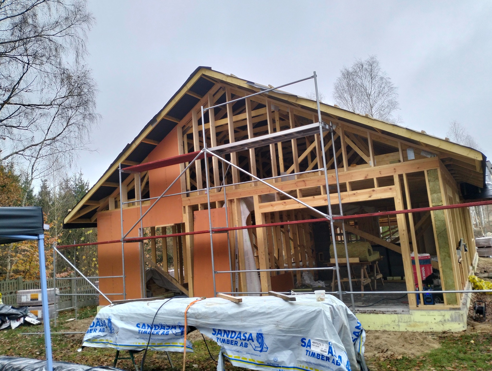 Budowa domu szkieletowego: drewniana konstrukcja, ściany z izolacją, rusztowania. Widoczny etap montażu, materiały budowlane i drzewa w tle. Pochmurne niebo.