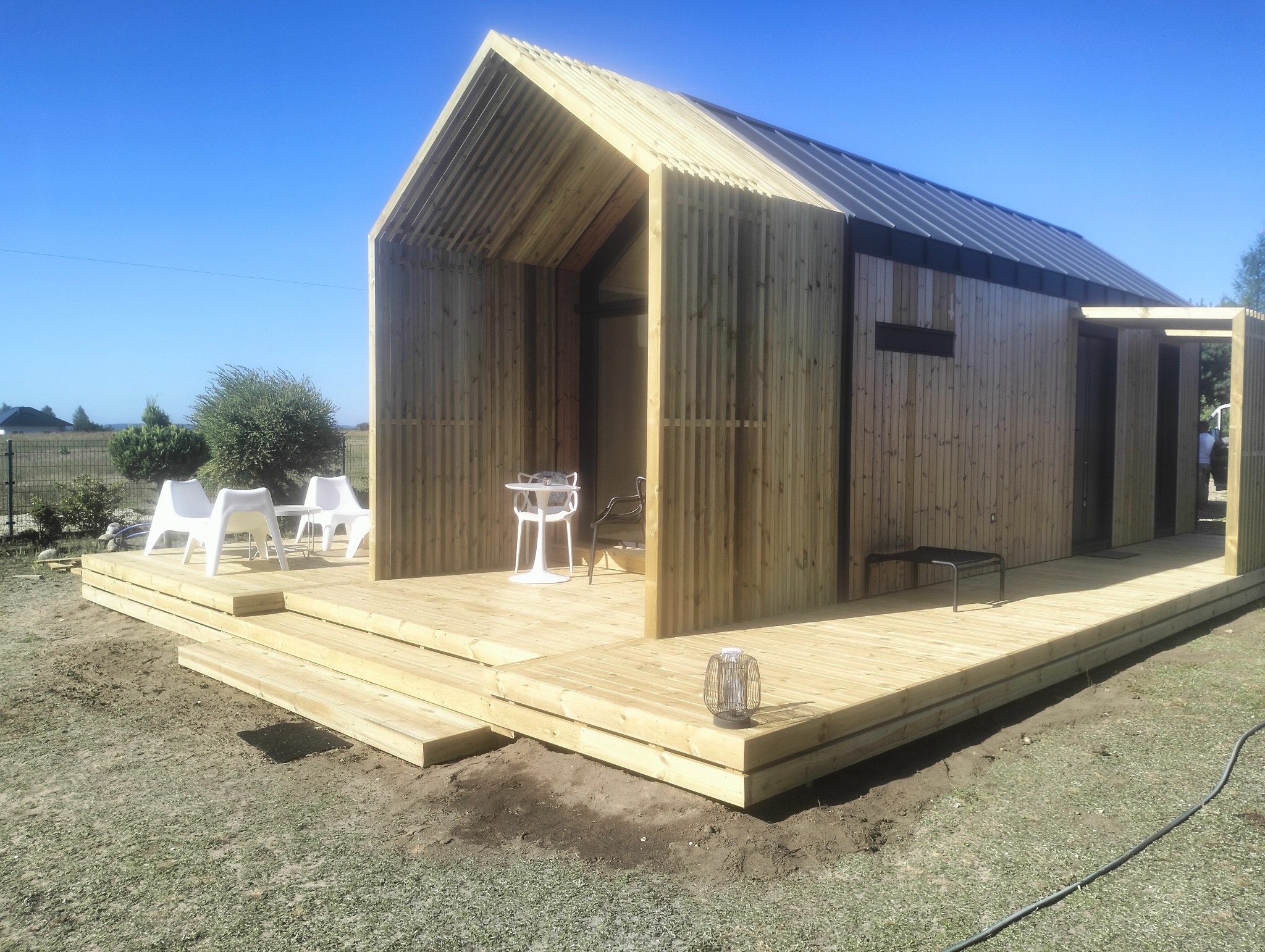 Nowoczesny dom szkieletowy z drewnianą elewacją i tarasem. Widoczne meble ogrodowe i oświetlenie zewnętrzne. Błękitne niebo w tle.