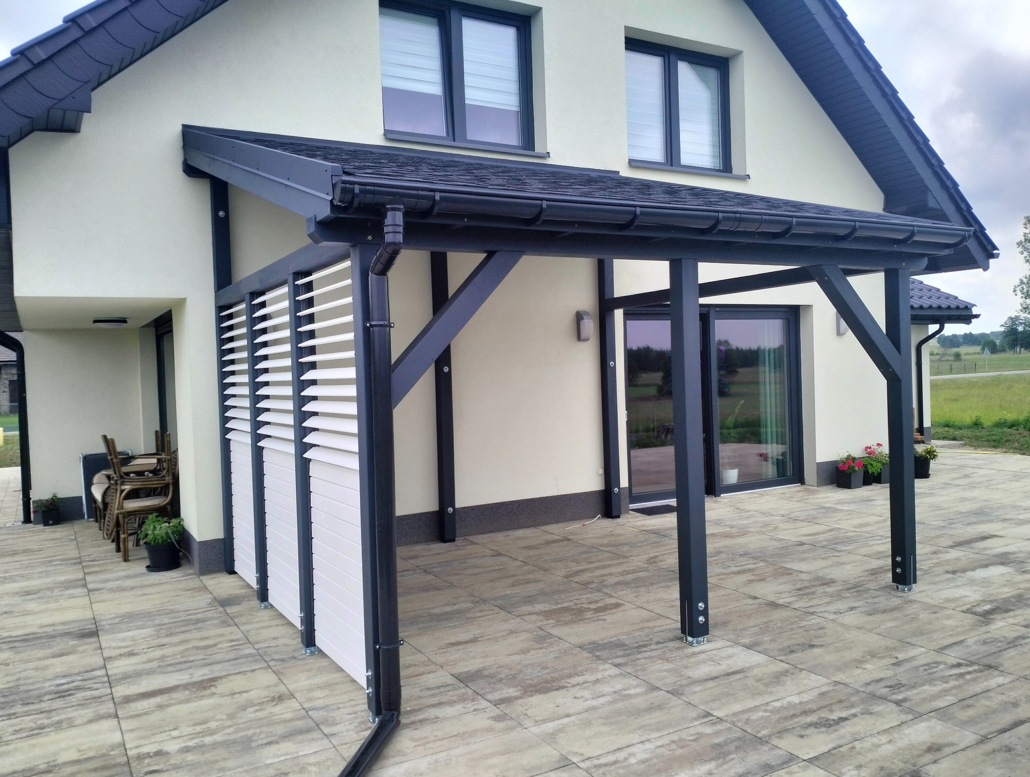 Nowoczesny dom z zadaszonym tarasem z białymi żaluzjami i ciemną konstrukcją. Widok na ogród i okna z roletami zewnętrznymi. Eleganckie wykończenie.