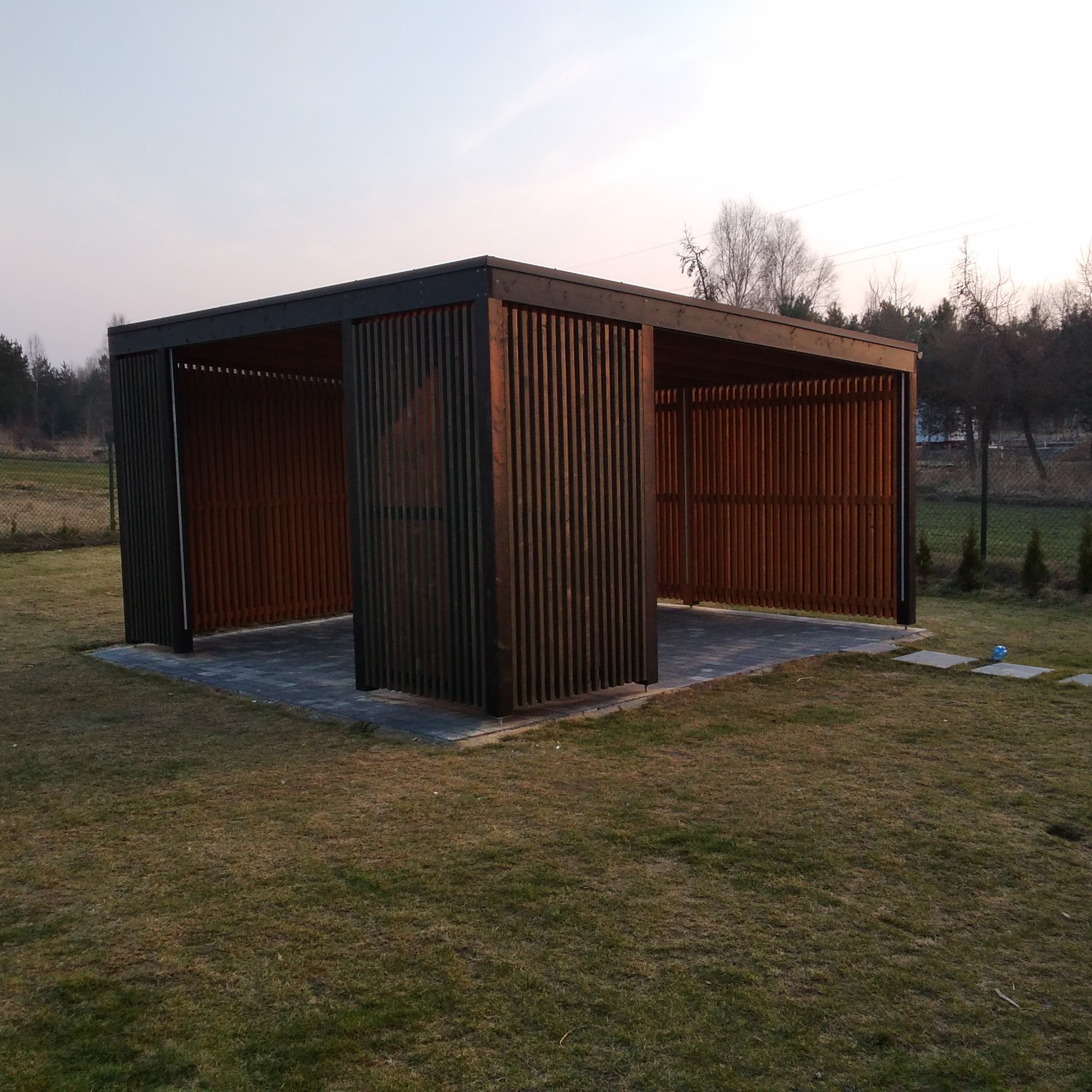 Nowoczesna, ażurowa altana ogrodowa z ciemnego drewna, zbudowana na kostce brukowej. W tle zieleń ogrodu i drzewa. Widok z zewnątrz, w świetle zachodzącego słońca.